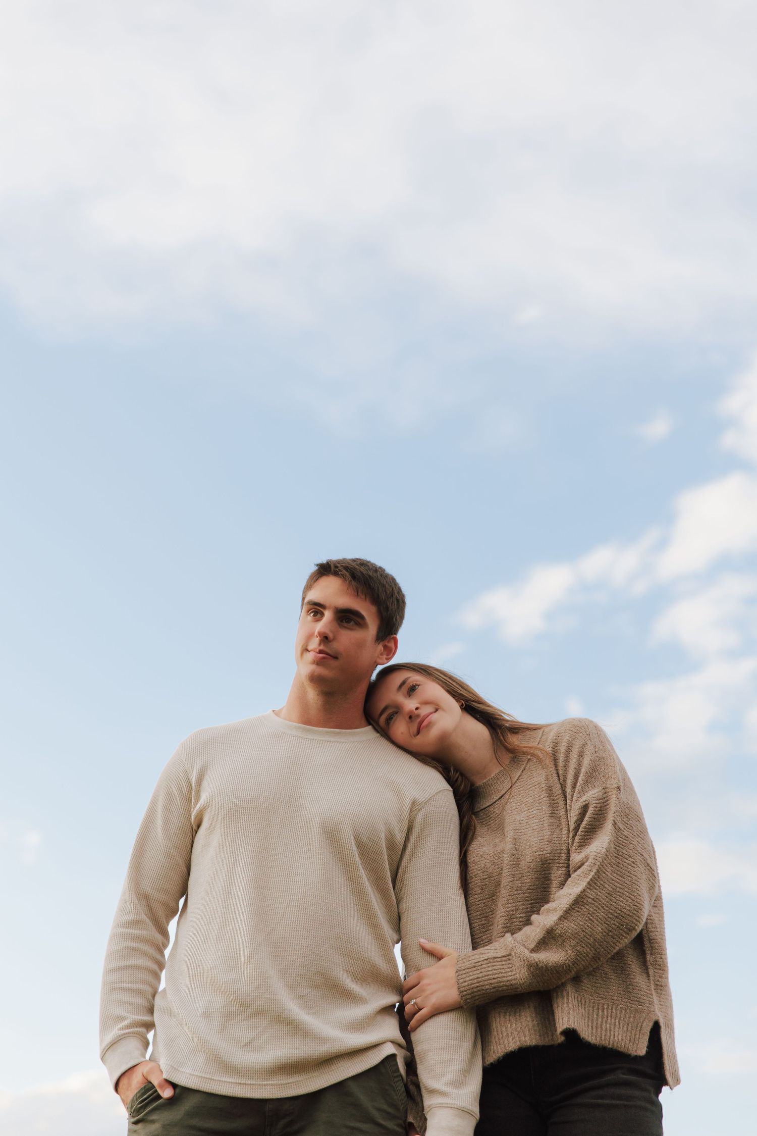 Couple in neutral colored sweaters poses against blue sky in romantic outdoor portraits.
