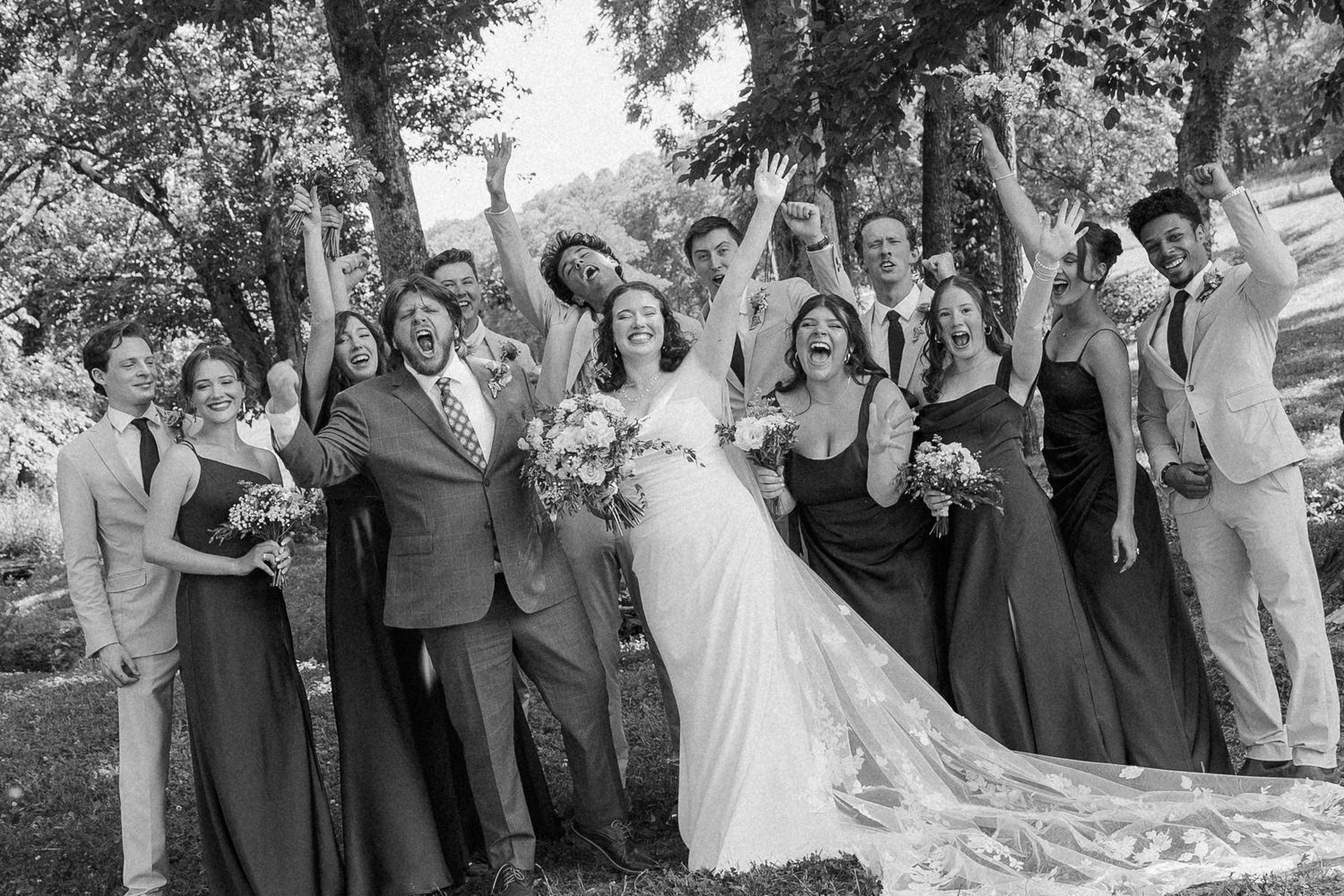 Black and white wedding party photo with bride, groom, bridesmaids, and groomsmen celebrating outdoors under trees.