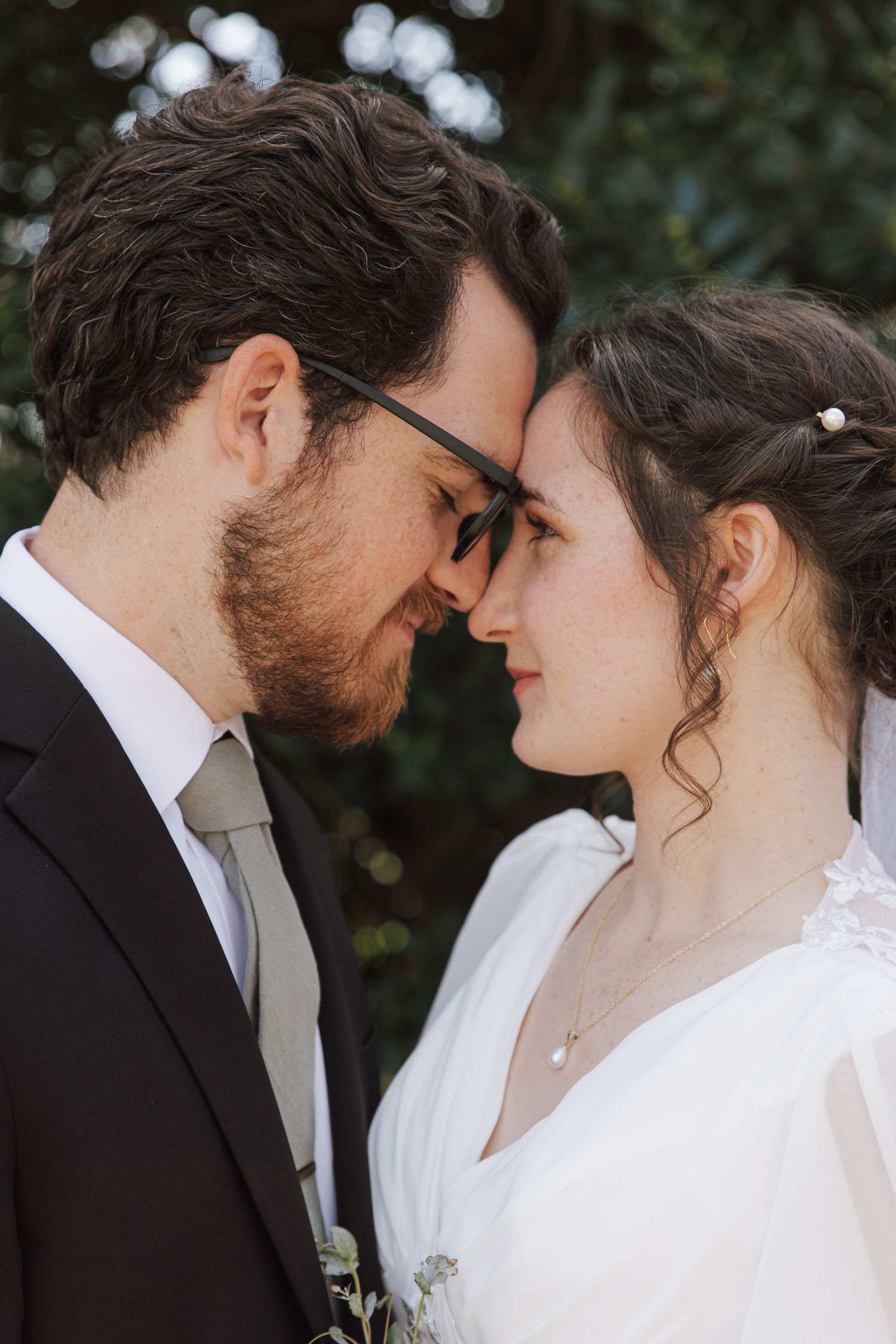 Bride and groom touching foreheads intimately, groom wearing glasses and suit, bride in white wedding dress outdoors.