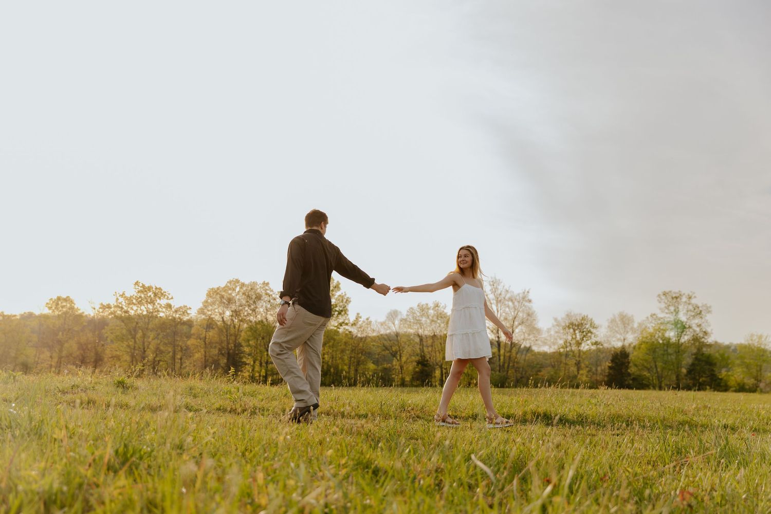 A couple dances together in a sunlit meadow during golden hour with trees in the background.