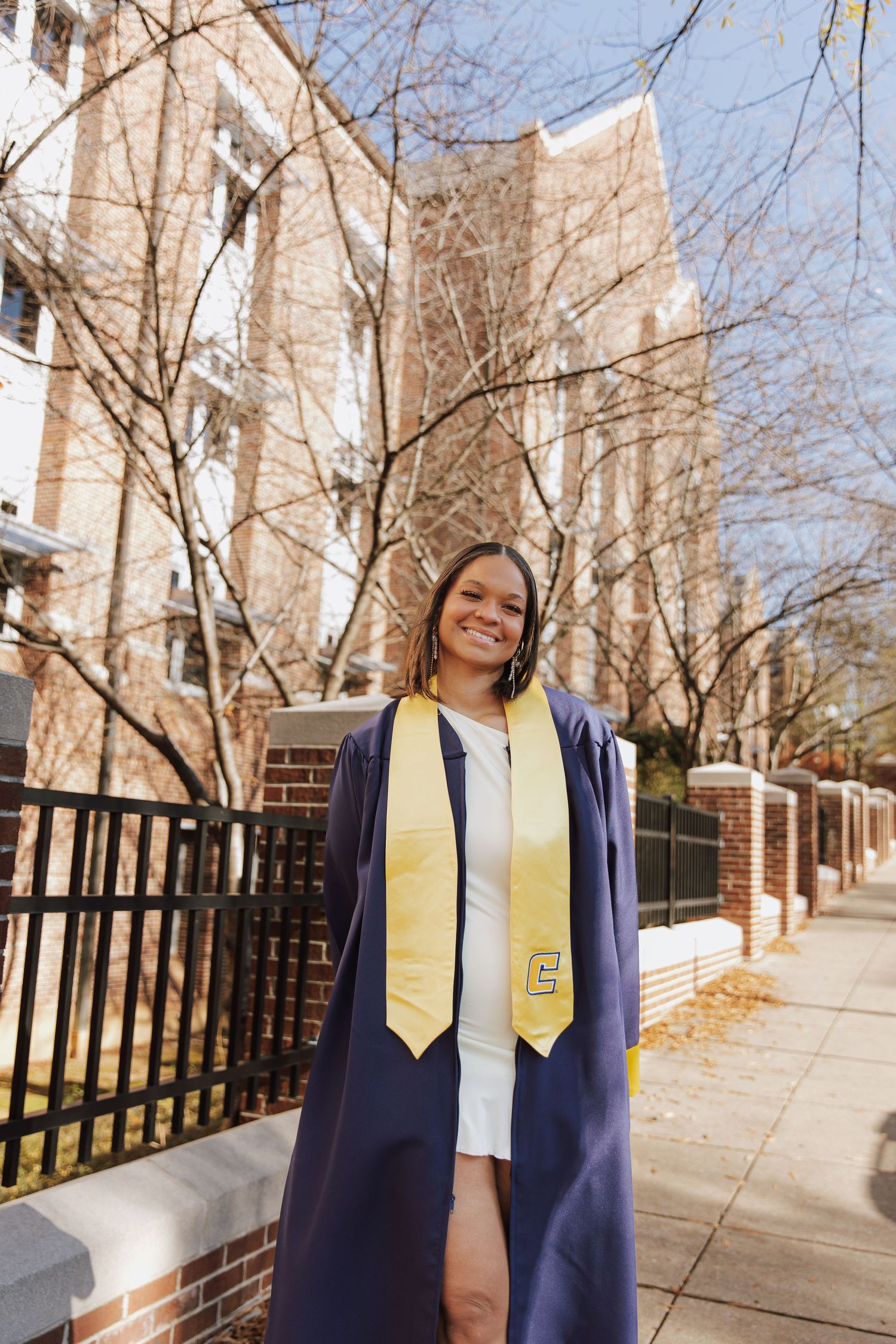 Graduate in navy gown and gold stole stands on campus walkway with brick buildings and fence.