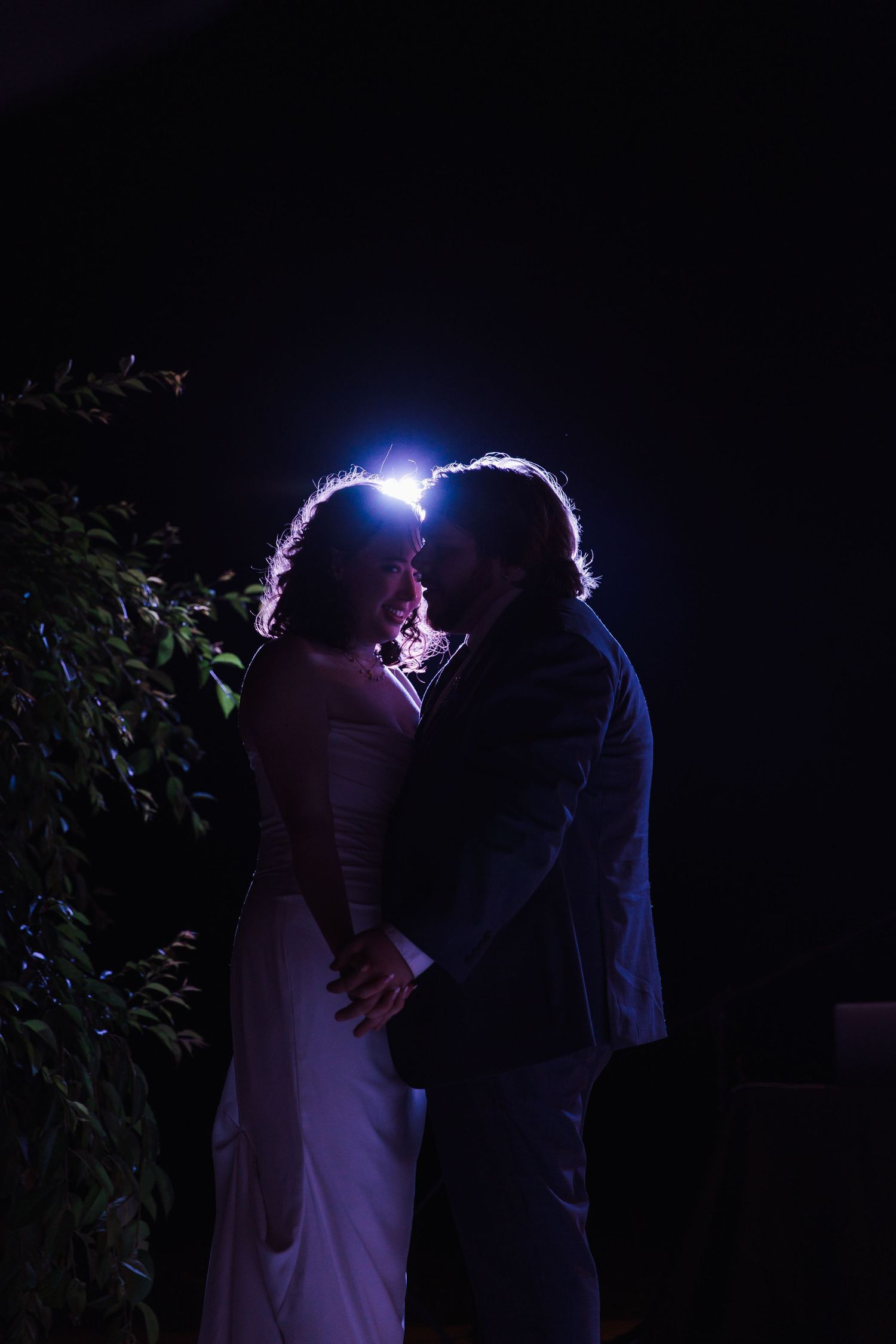 A bride and groom share a romantic kiss at night, illuminated by a soft backlight with lush greenery in the background.