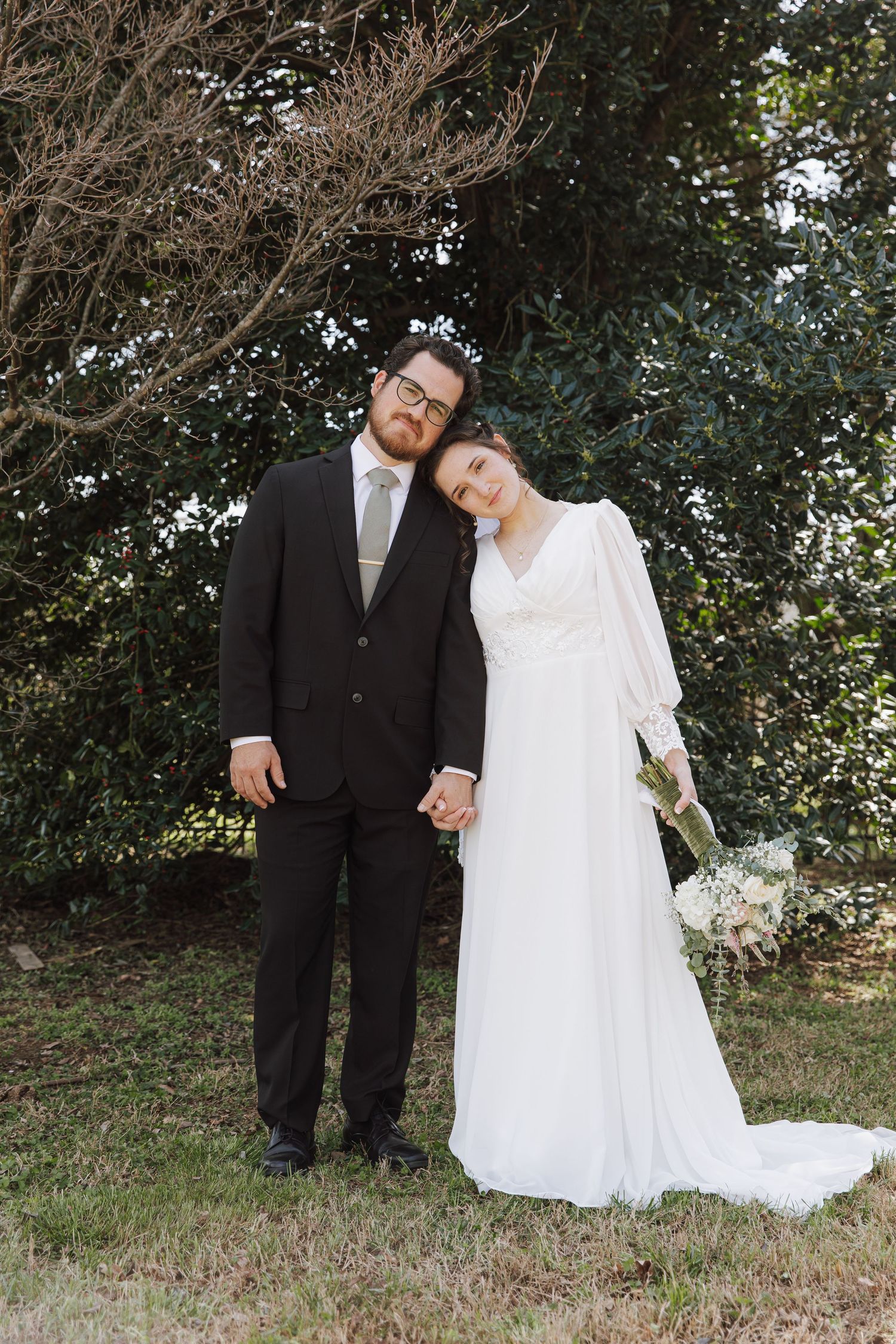 Bride leaning on groom's shoulder holding hands outdoors, she wears a long-sleeve white gown with bouquet.