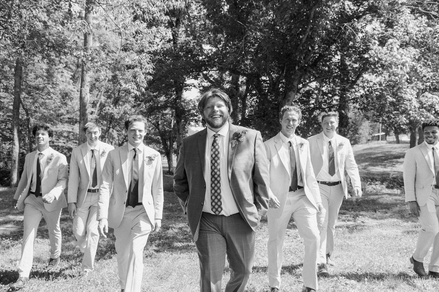 Groom and groomsmen walk confidently together in suits through a tree-lined outdoor wedding venue.