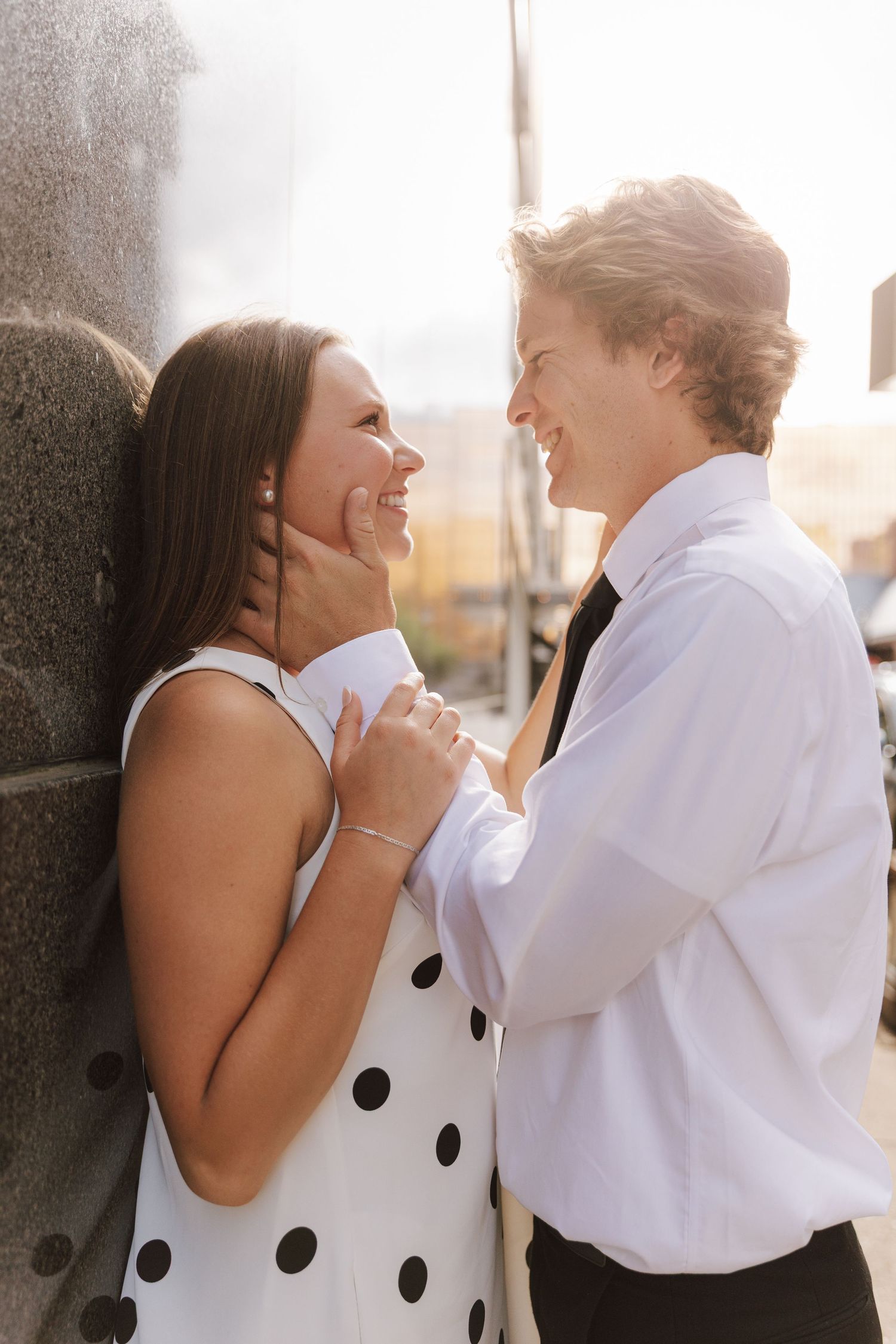 A couple shares a joyful moment together against a wall, with her wearing a polka dot dress and him in a white shirt.