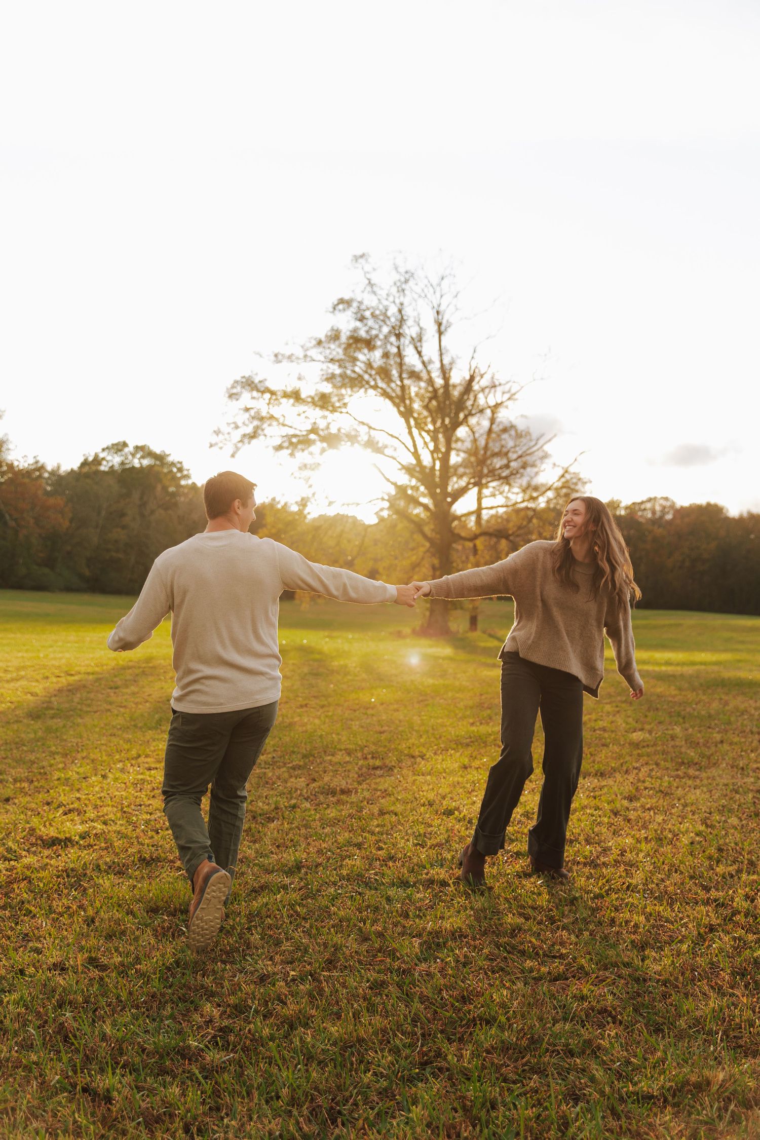 Couple holds hands while spinning playfully in a sunlit field during golden hour.