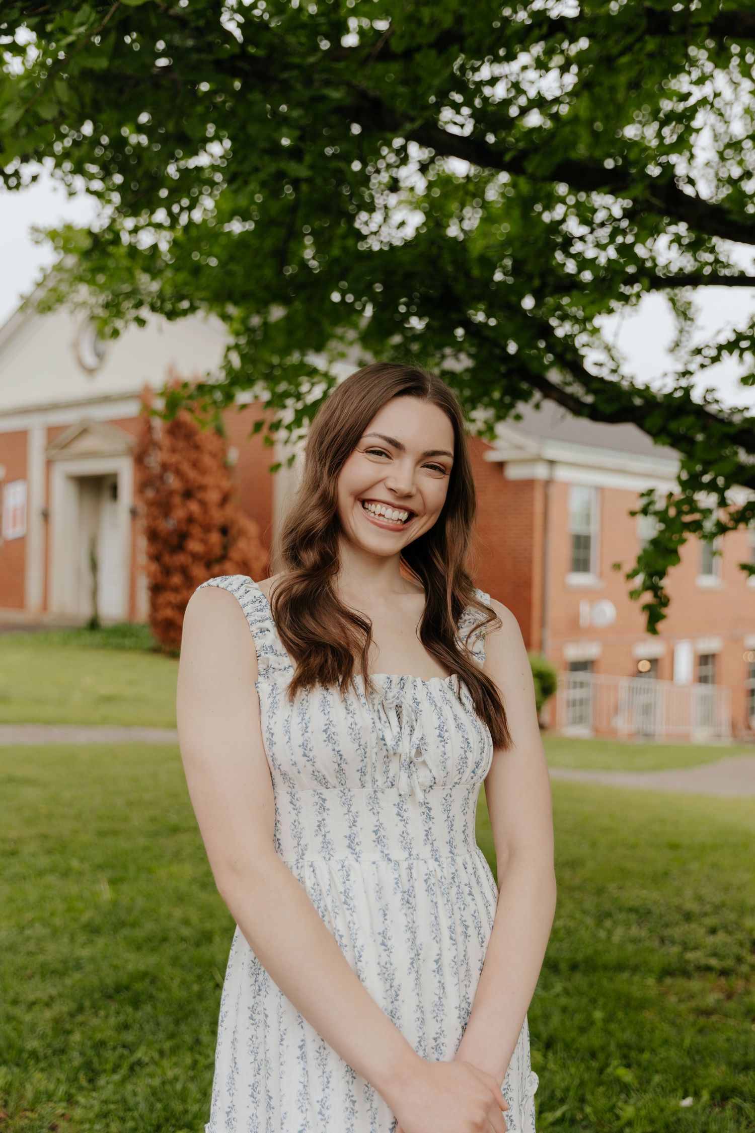 A person in a white lace dress smiles while sitting on grass with brick buildings visible in the background.
