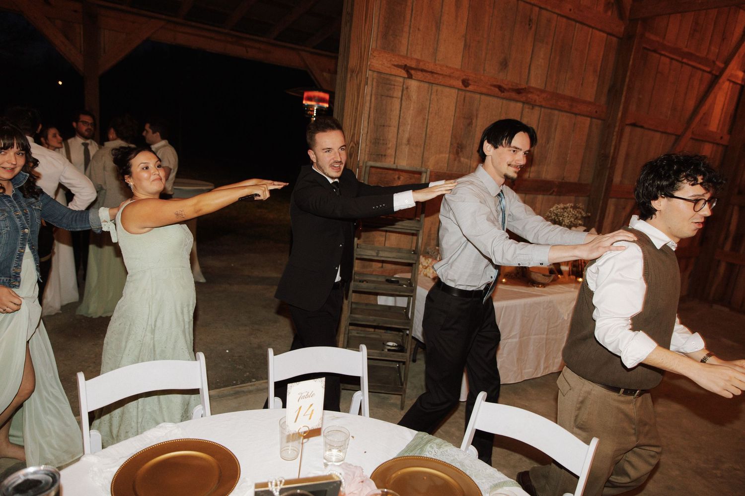 Wedding guests dancing in a chain line at rustic barn reception venue during evening celebration.