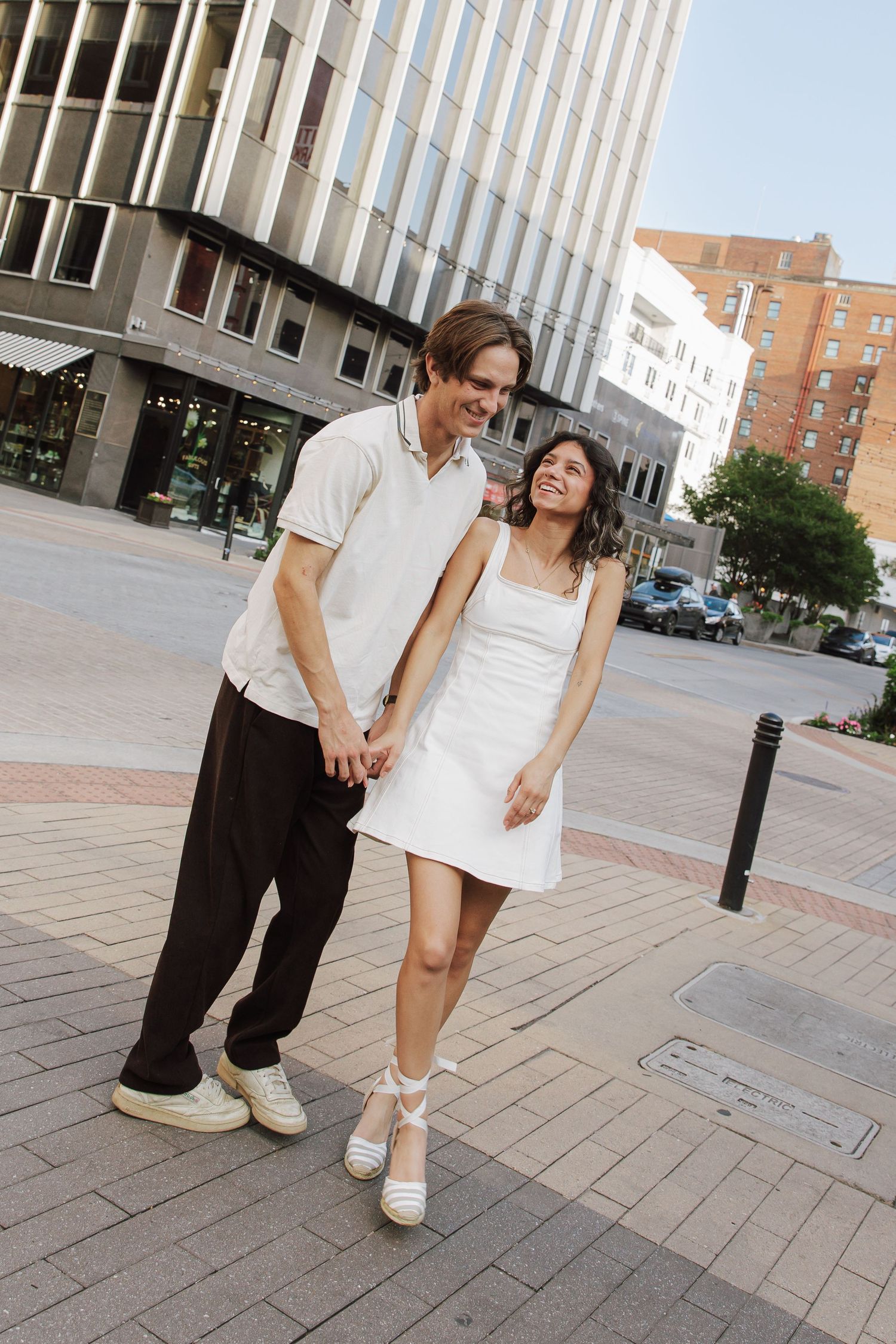 Happy couple holding hands walking through urban city street during outdoor engagement photoshoot.