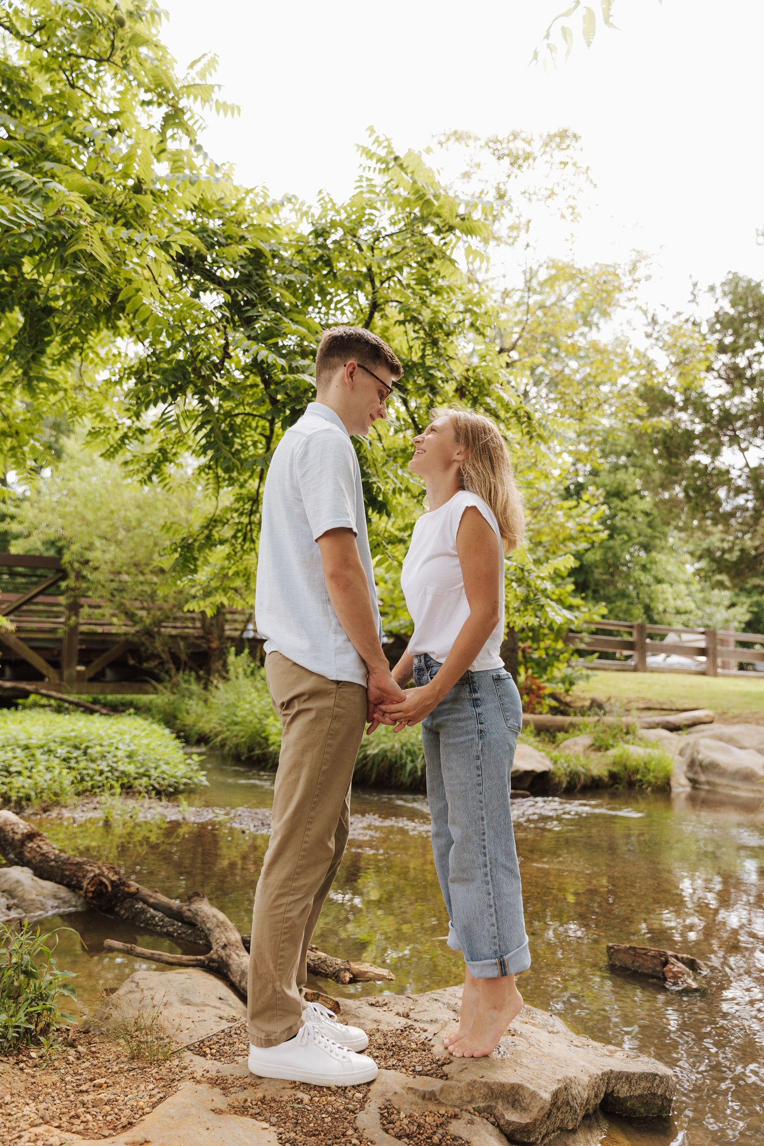 A couple stands together on rocks near a peaceful stream surrounded by lush green trees on a sunny day.