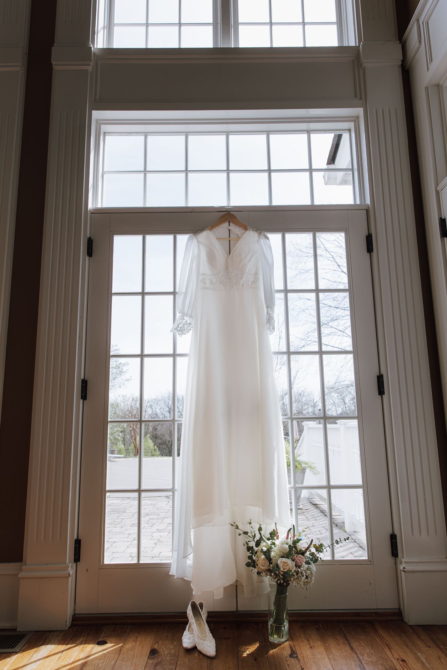 White wedding dress hanging in window with bridal bouquet and heels below.