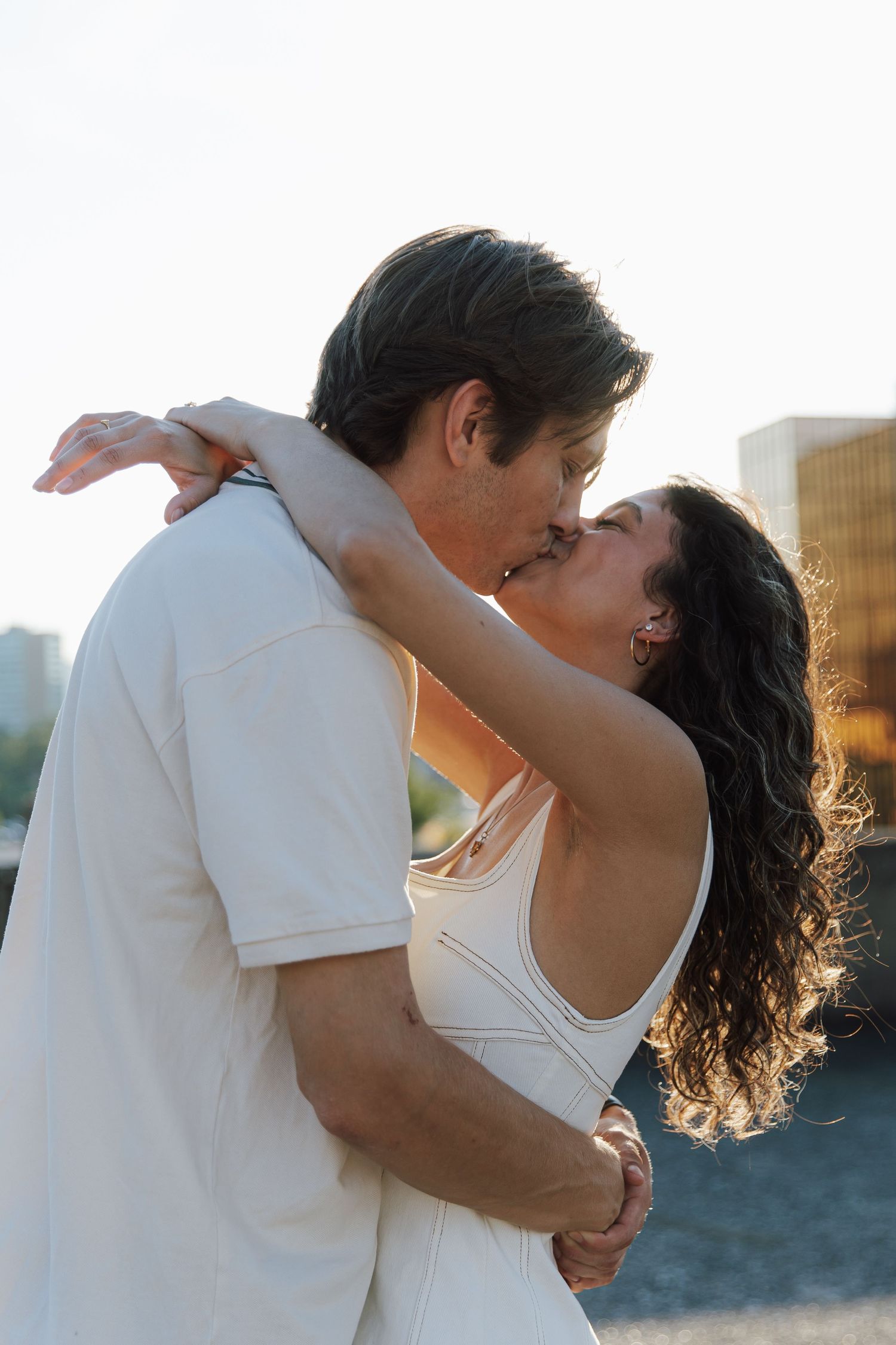 Romantic couple sharing a tender kiss outdoors at golden hour, embracing against a soft city backdrop.