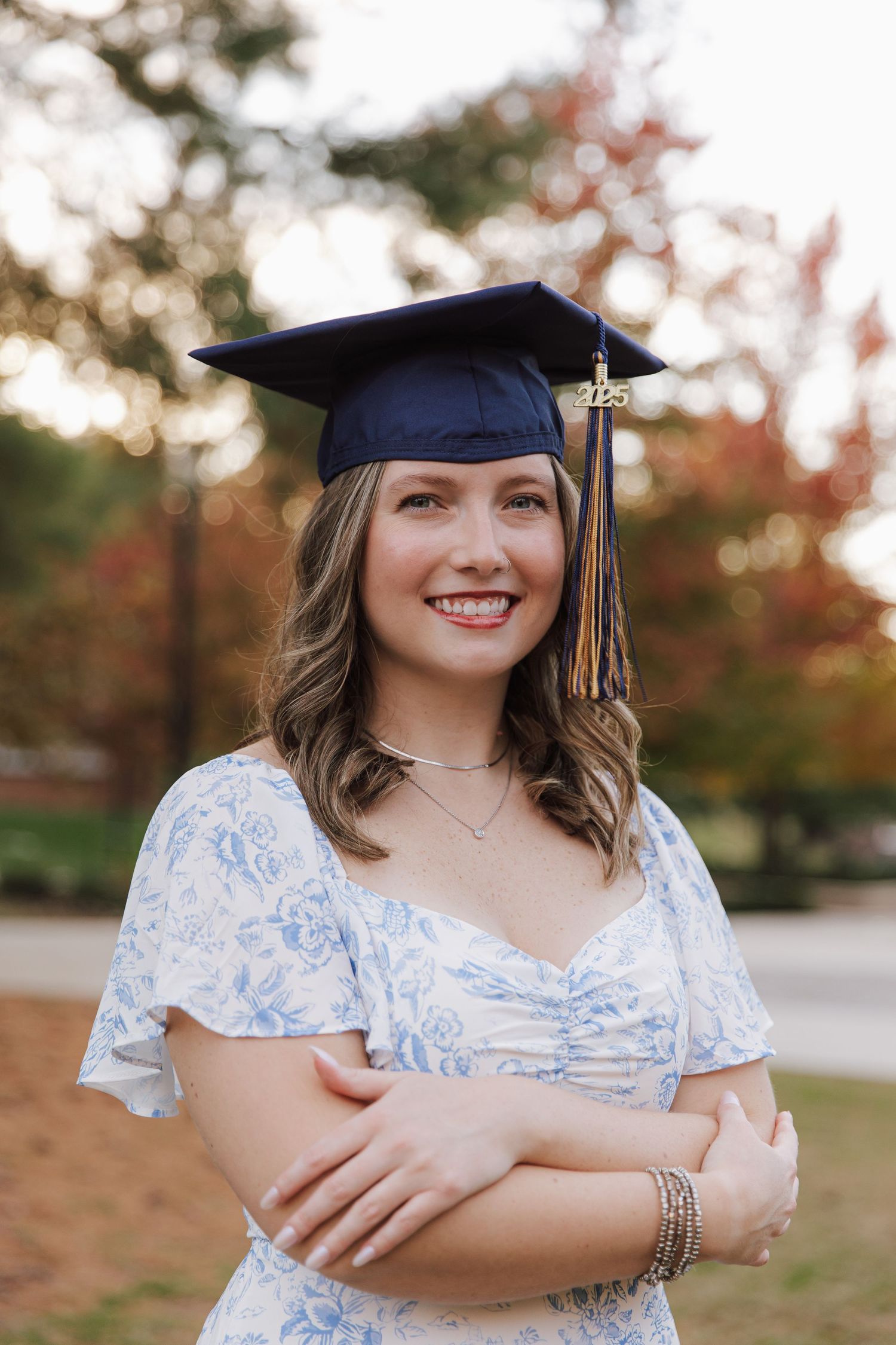 Graduate in blue and white floral dress poses outdoors with graduation cap and tassel during autumn season.