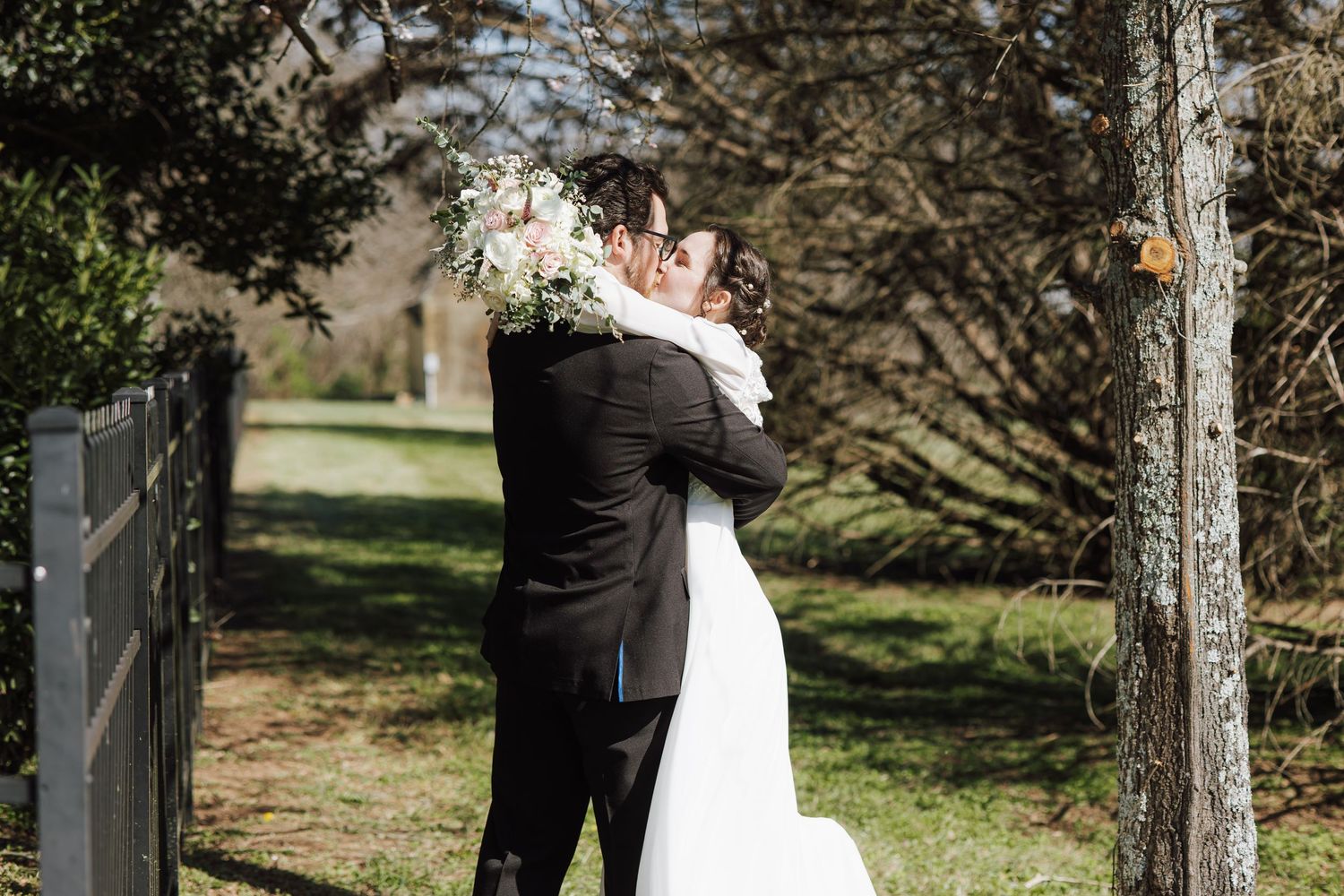 Bride and groom embracing outdoors on a sunny day, surrounded by lush green grass and trees at a wedding venue.