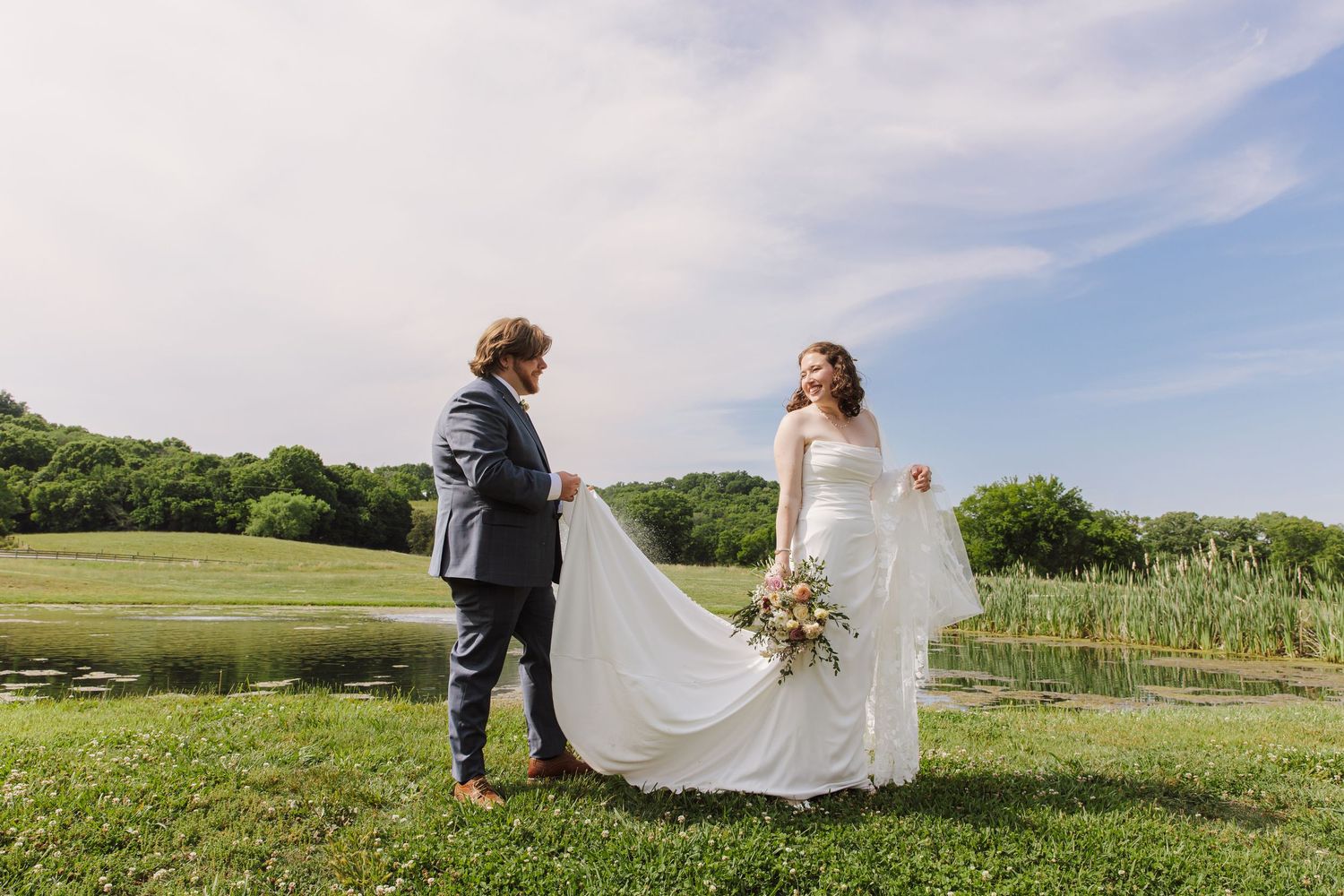 Bride and groom stand by a pond, groom holding her white veil as she smiles under a blue sky.
