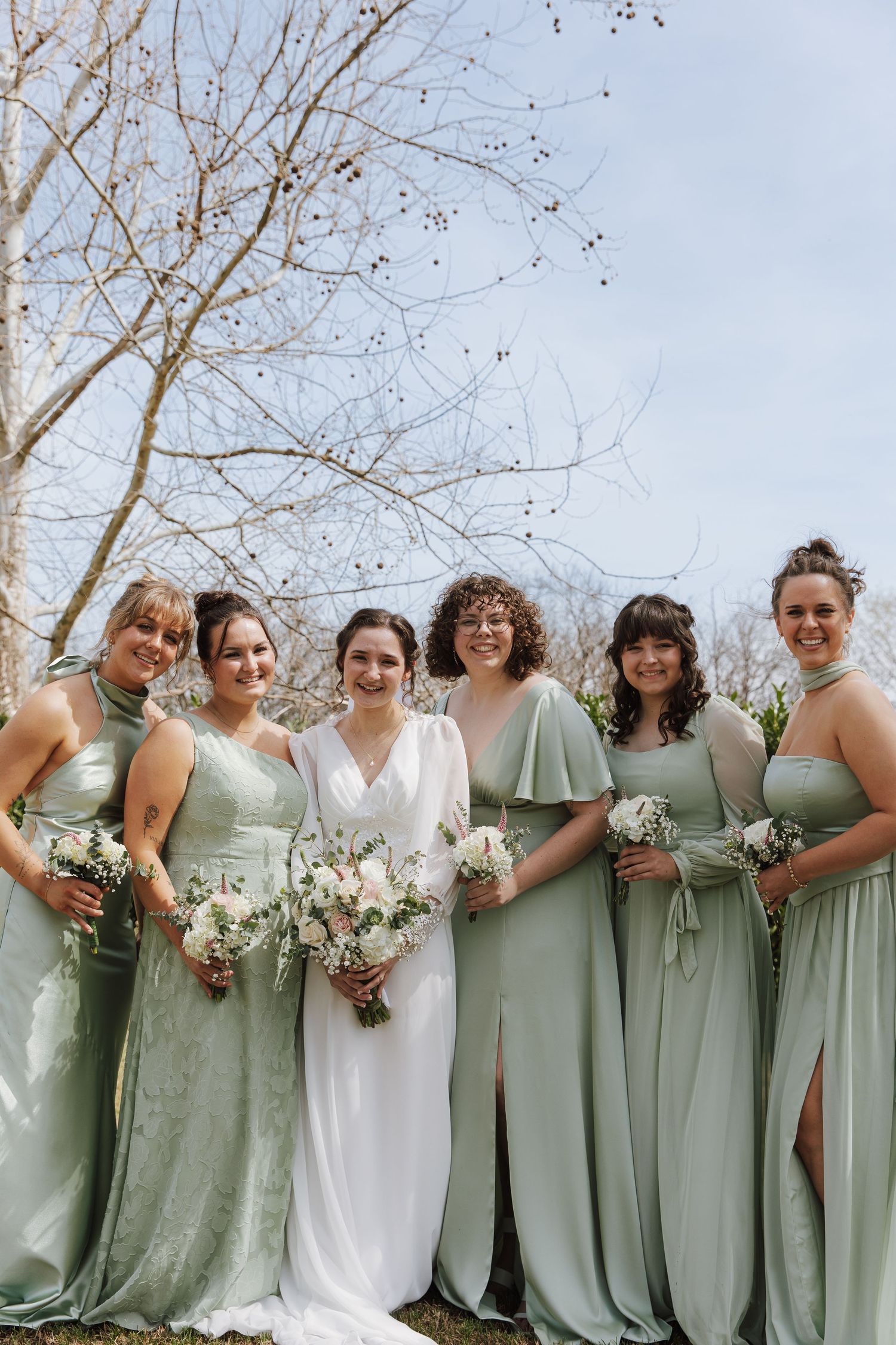 Bridesmaids in sage green dresses stand together holding white bouquets against a winter backdrop.