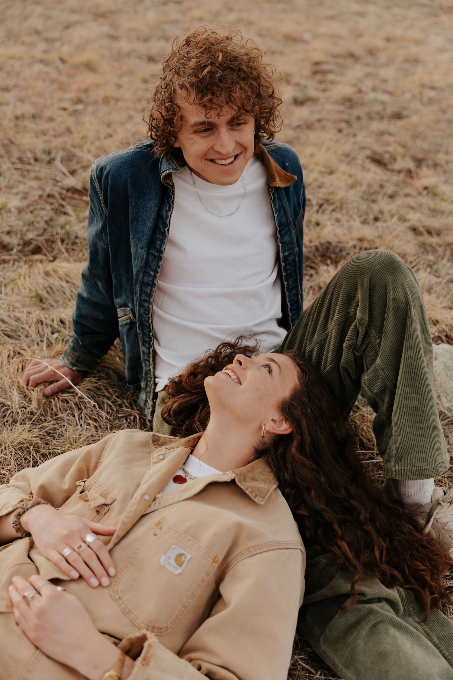 Two people relaxing in a casual autumn setting on dry grass wearing denim and neutral colored clothing.