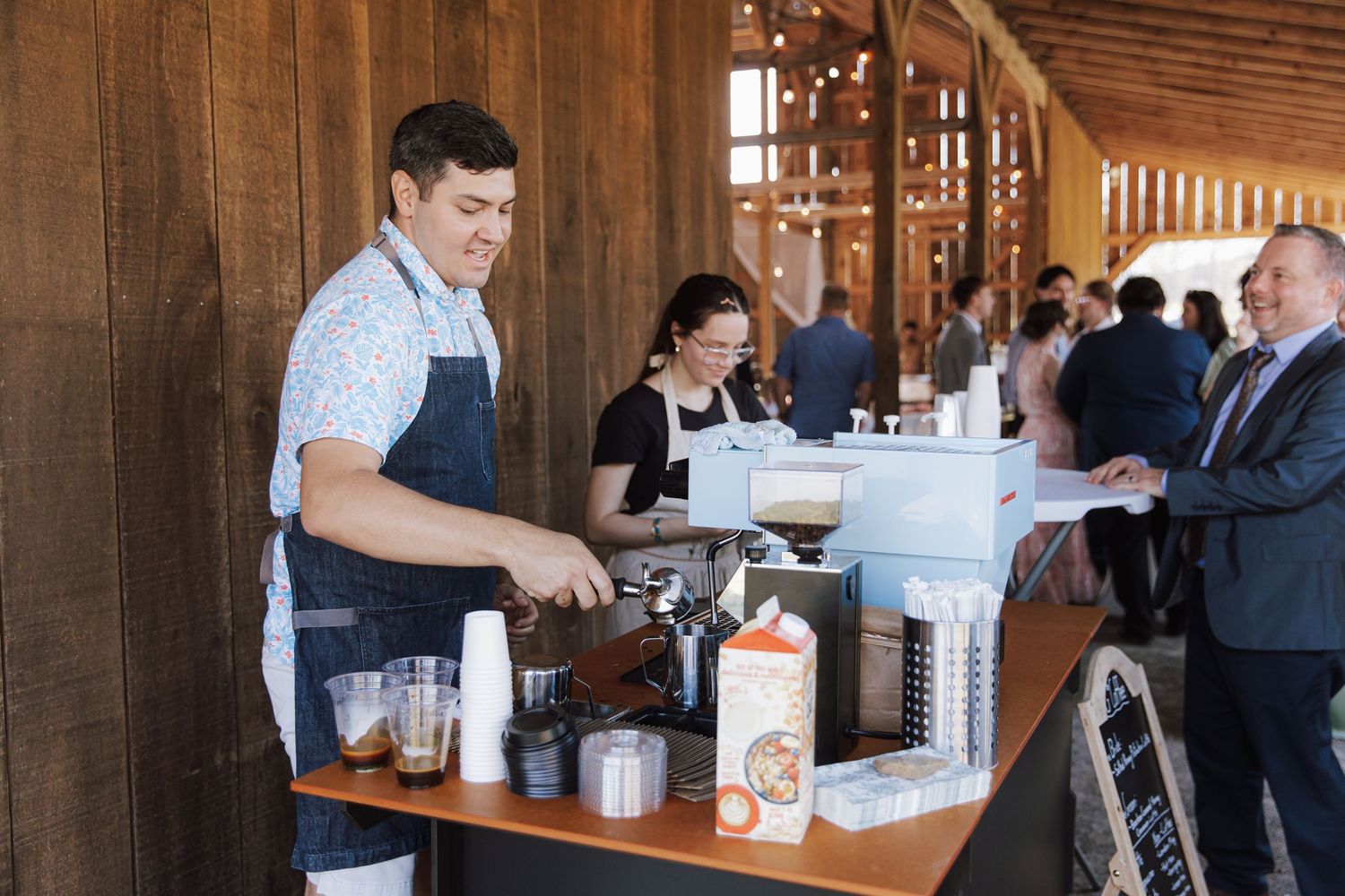 Barista in apron serves coffee at a wooden bar during a catered event with guests in background.