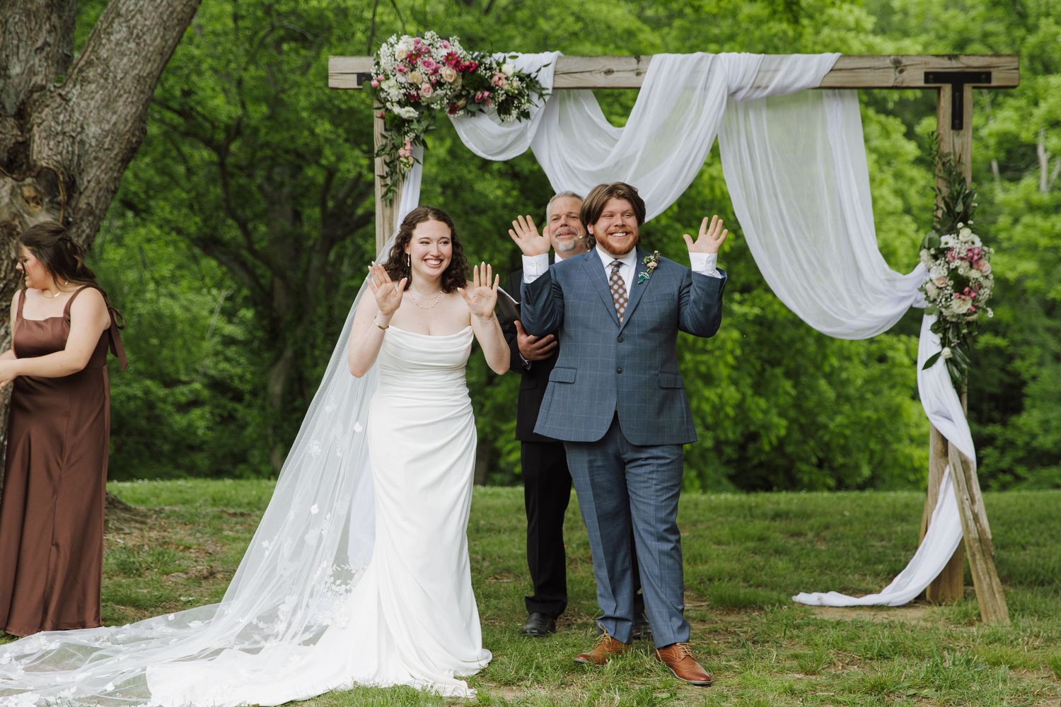 Joyful newlyweds celebrate under a rustic floral arch at an outdoor wedding ceremony on a lush green lawn.