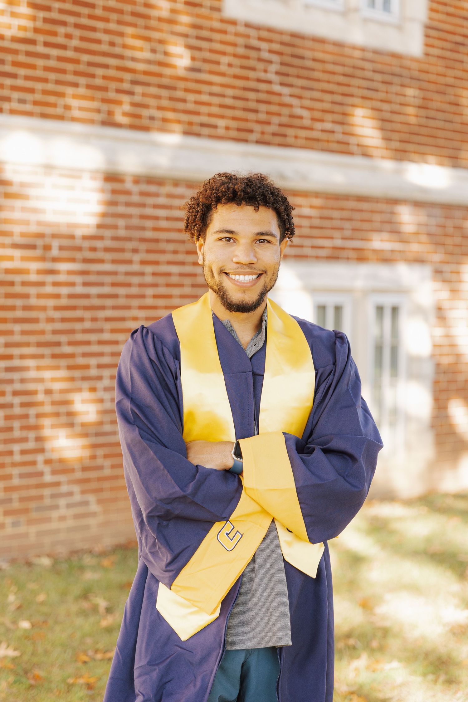Graduate in navy blue cap and gown with gold stole poses joyfully against brick wall in outdoor setting.