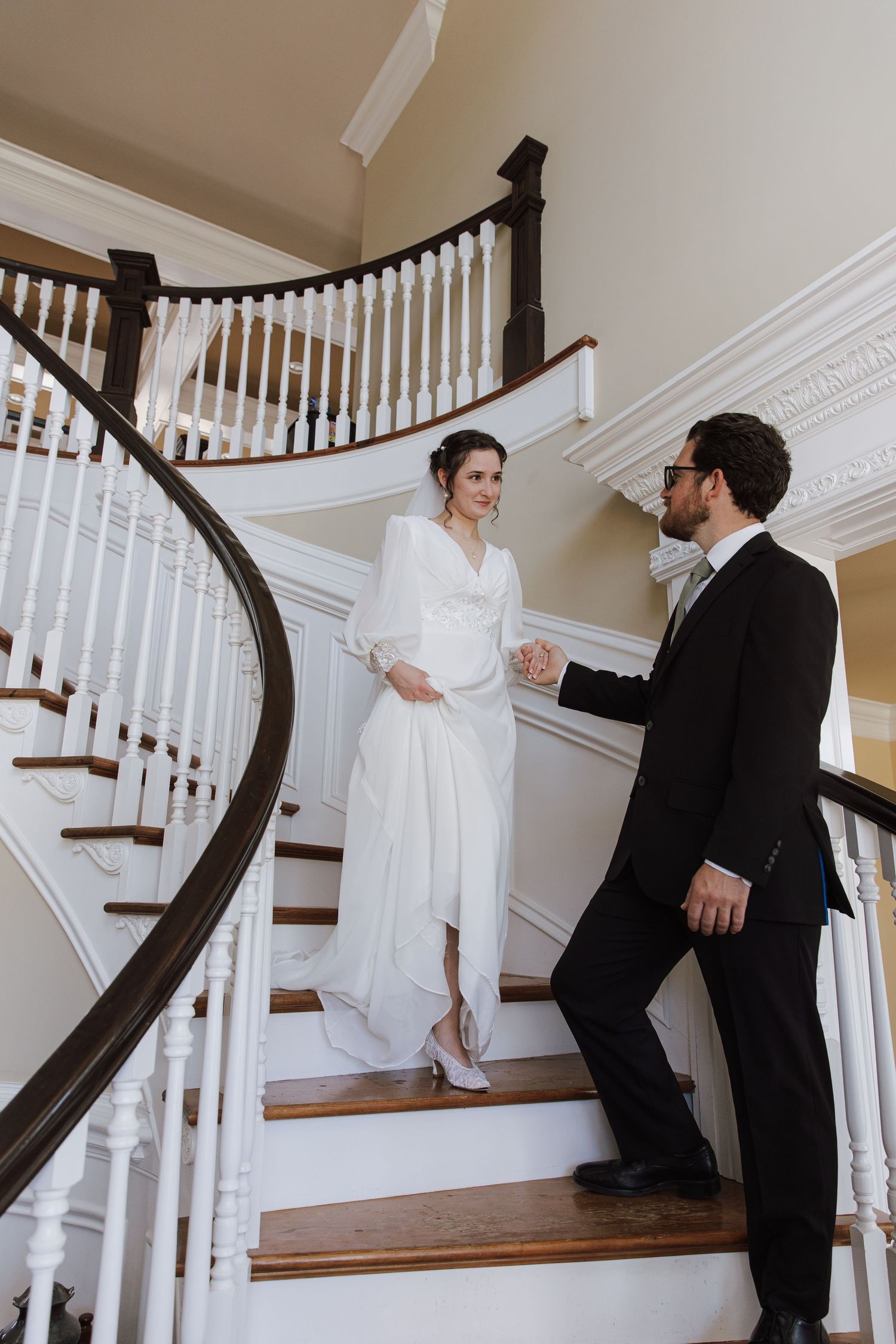Bride and groom holding hands on elegant curved staircase, bride in long-sleeve white wedding dress smiling.
