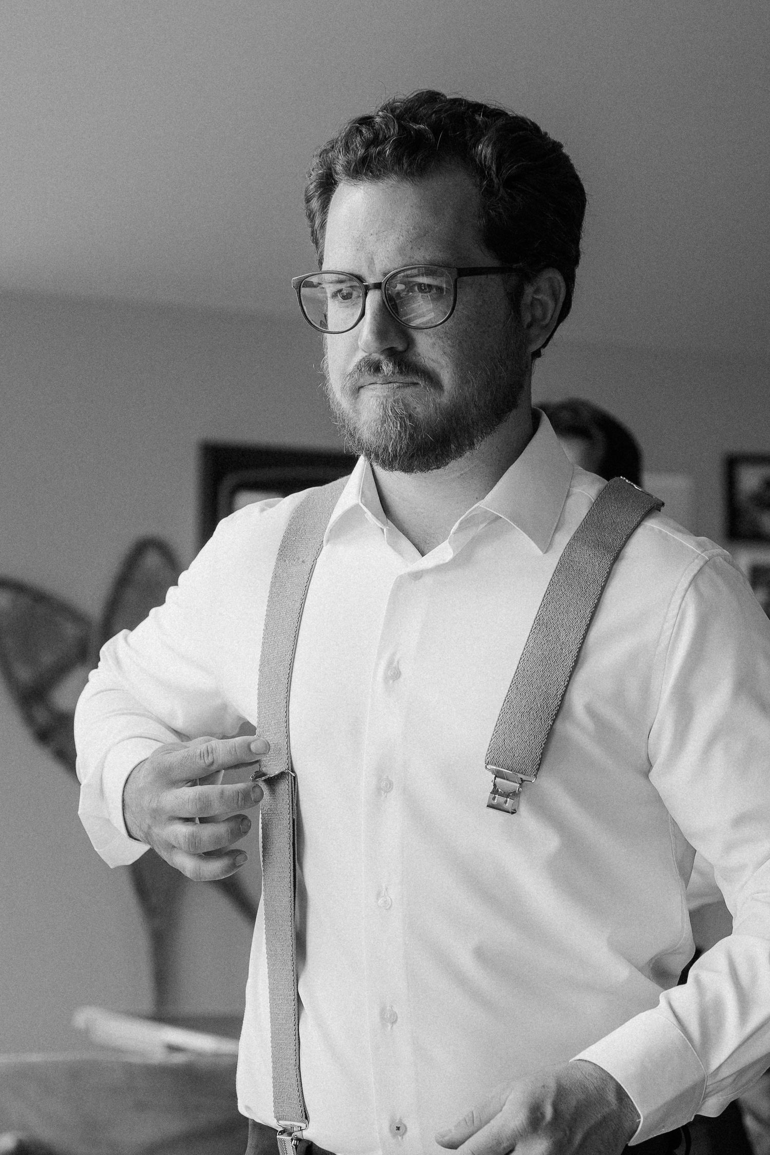 Black and white portrait of a bearded groom in white shirt and suspenders adjusting his tie indoors.