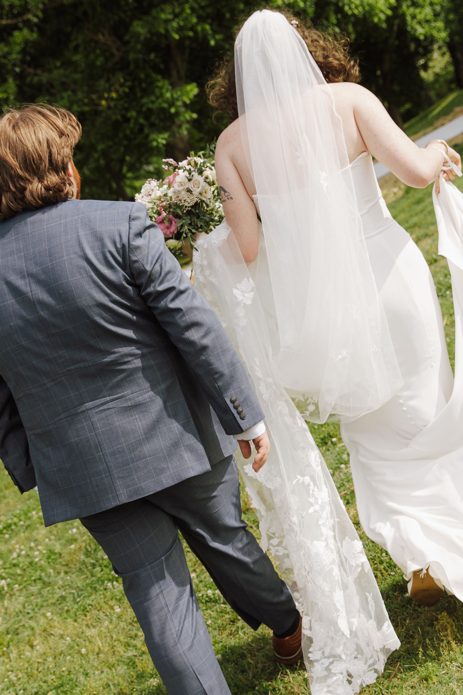 Bride in white lace gown and veil walks with groom in gray suit across green lawn holding bouquet.