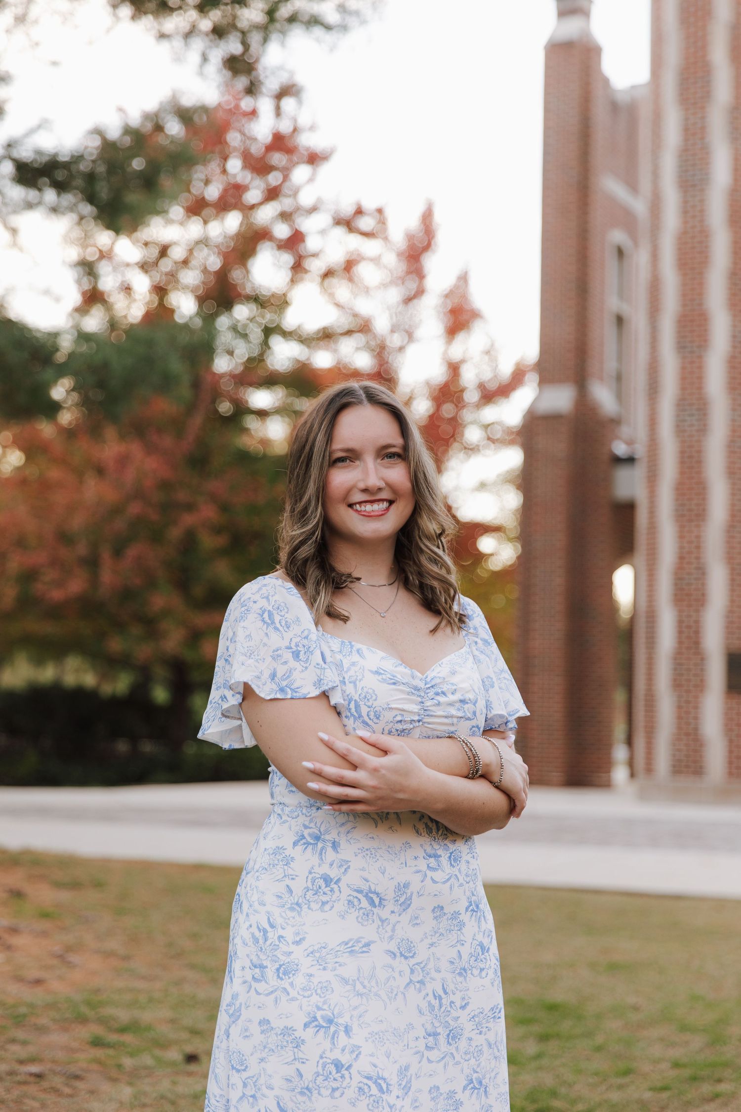 Graduate in blue and white floral dress poses outdoors near brick columns during sunset.