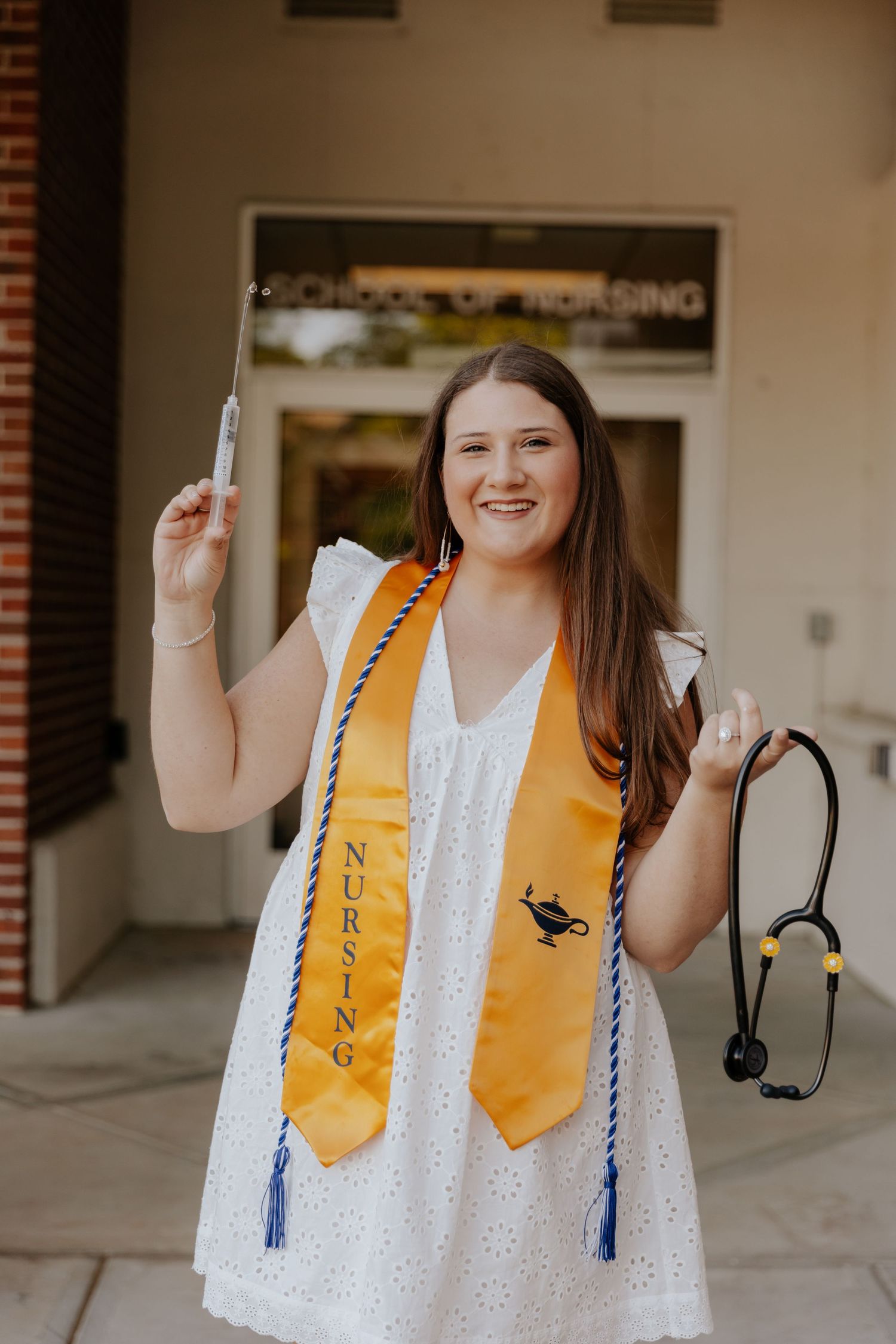 A nursing graduate in white dress holds stethoscope and displays nursing graduation stole.