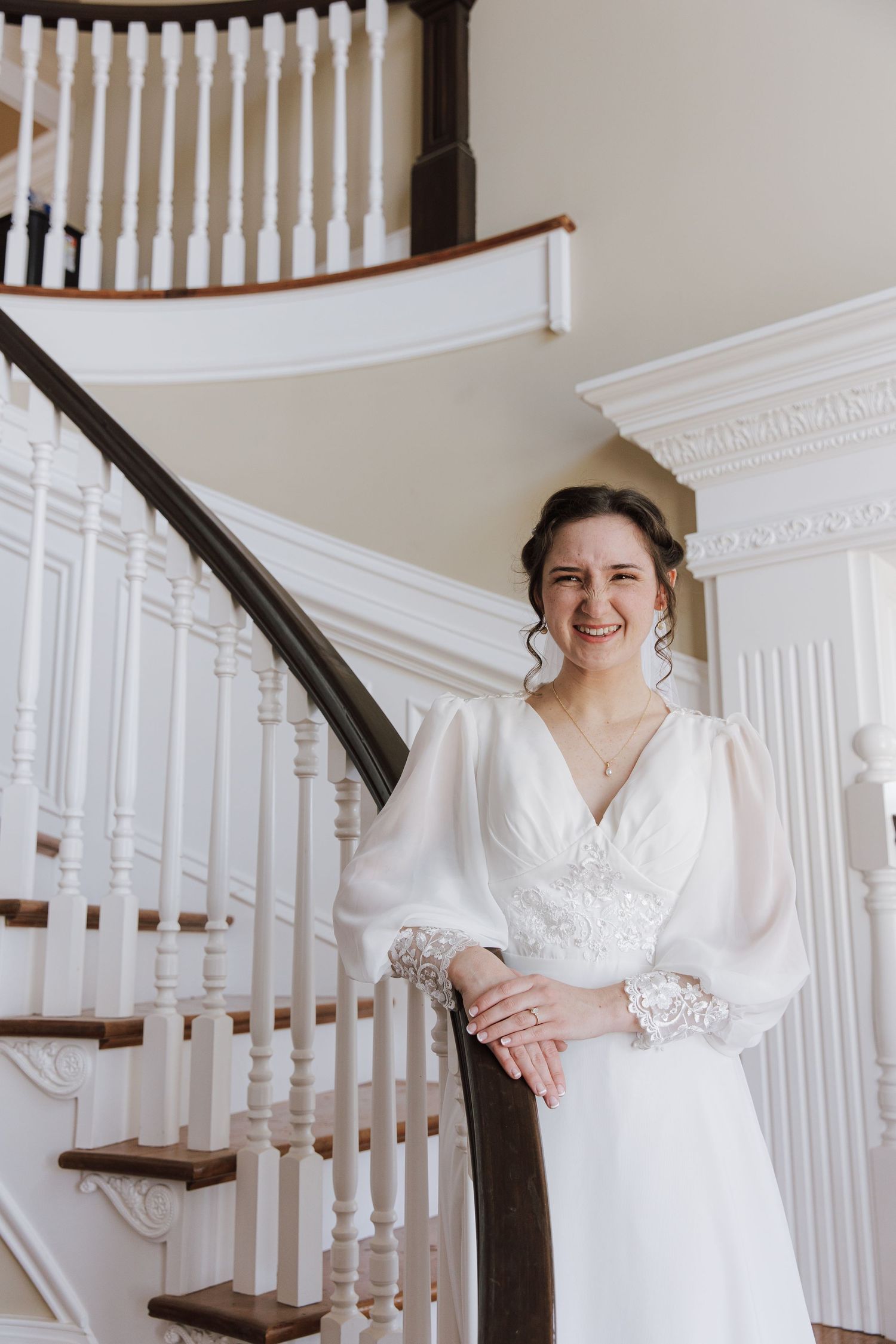 Smiling bride in white gown with long sleeves poses by elegant staircase in a classic white interior.