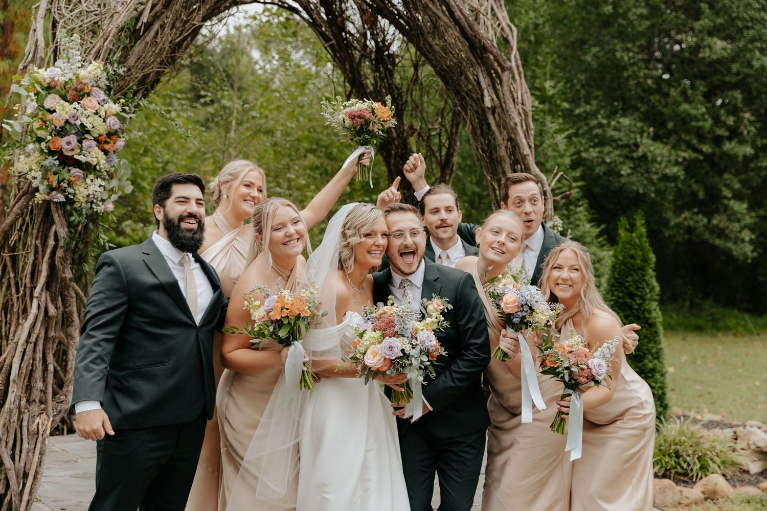 Wedding party poses outdoors in a garden setting with bridesmaids in champagne dresses.