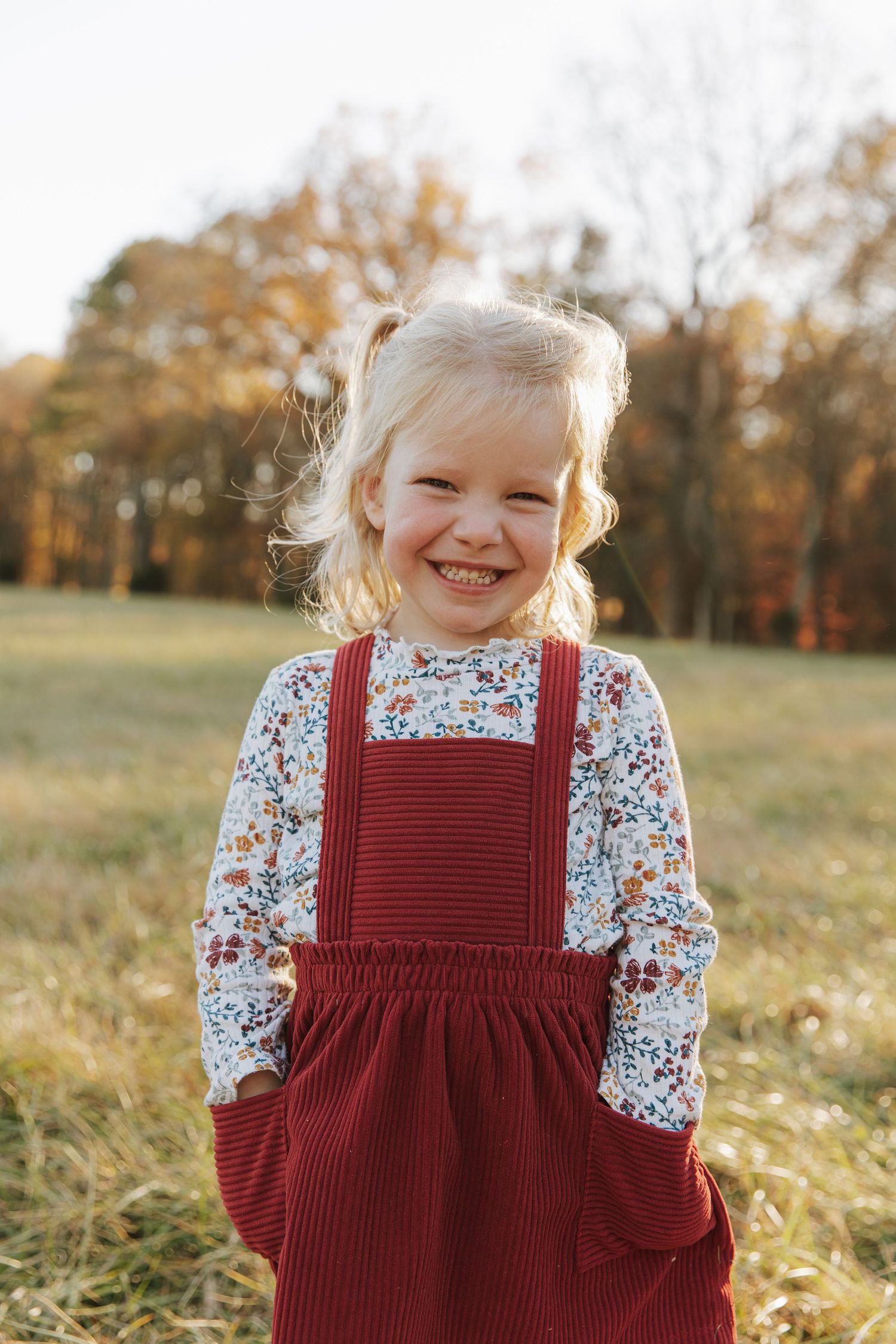 A child wearing a burgundy corduroy jumper and floral top plays in a golden autumn field at sunset.