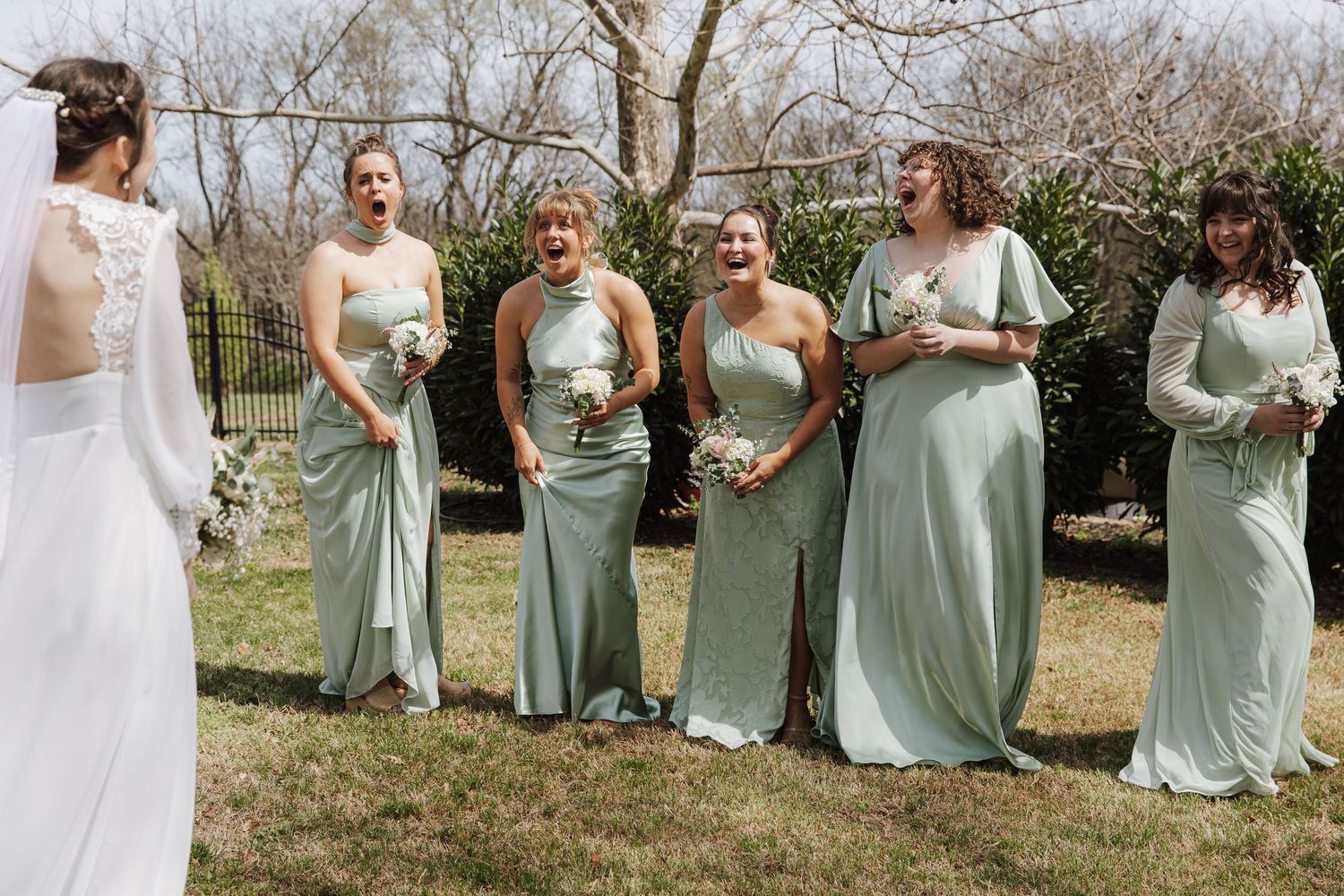 Bridesmaids in sage green gowns react with joy seeing the bride in a white dress outdoors at a spring wedding.