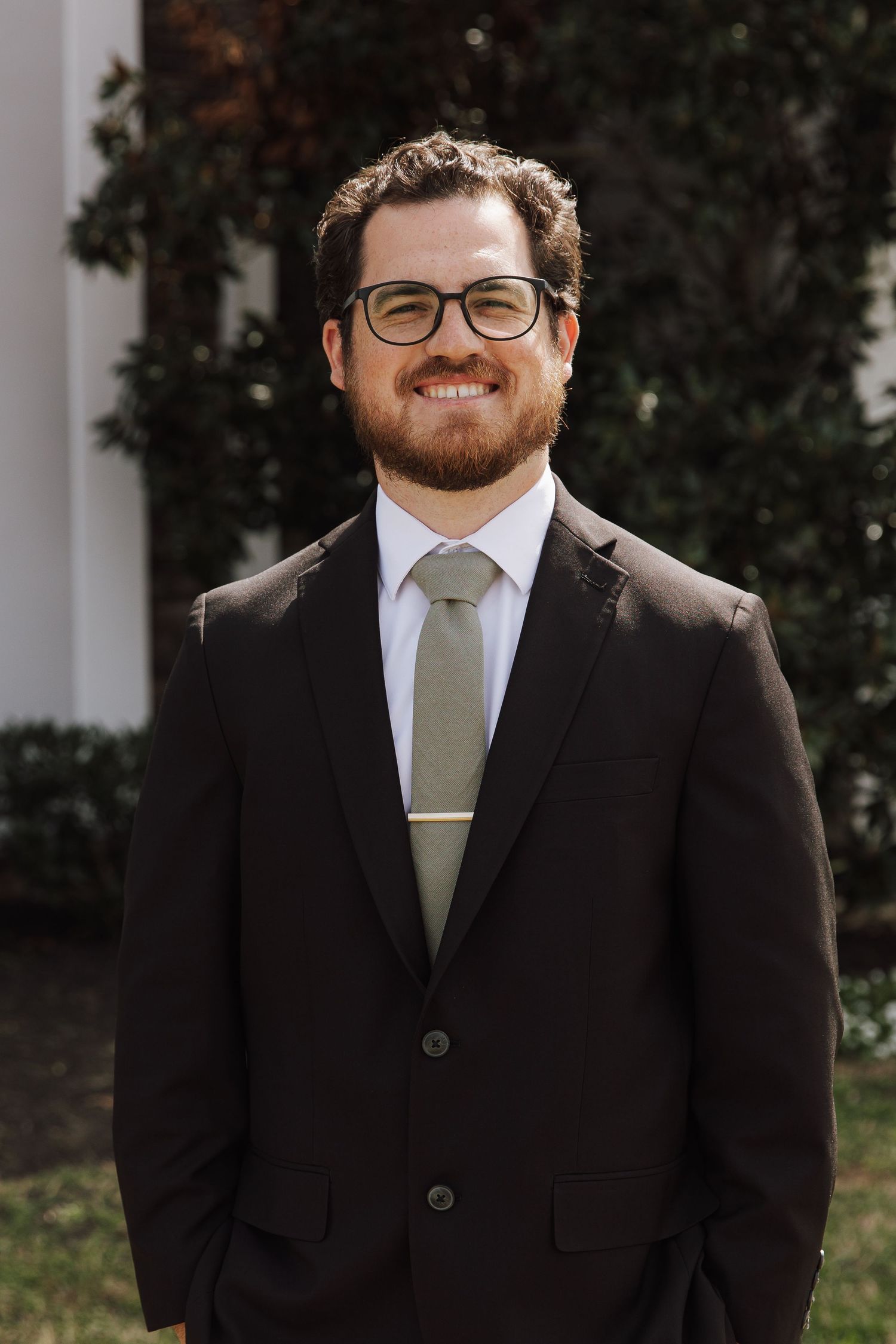 Smiling man with beard and glasses wearing dark suit and sage green tie outdoors.