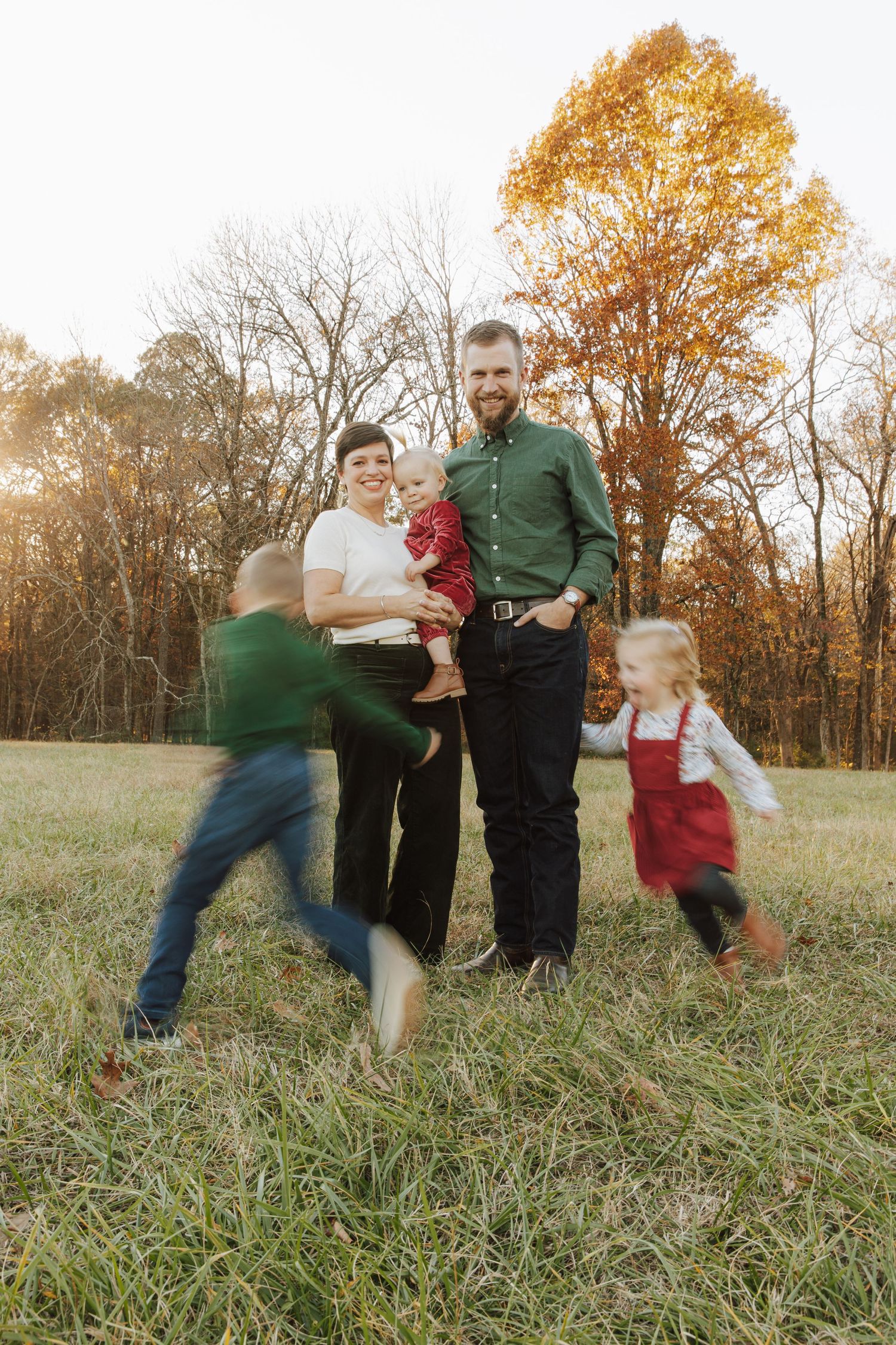 A family poses in a field during autumn with blurred motion of children running, set against golden trees.