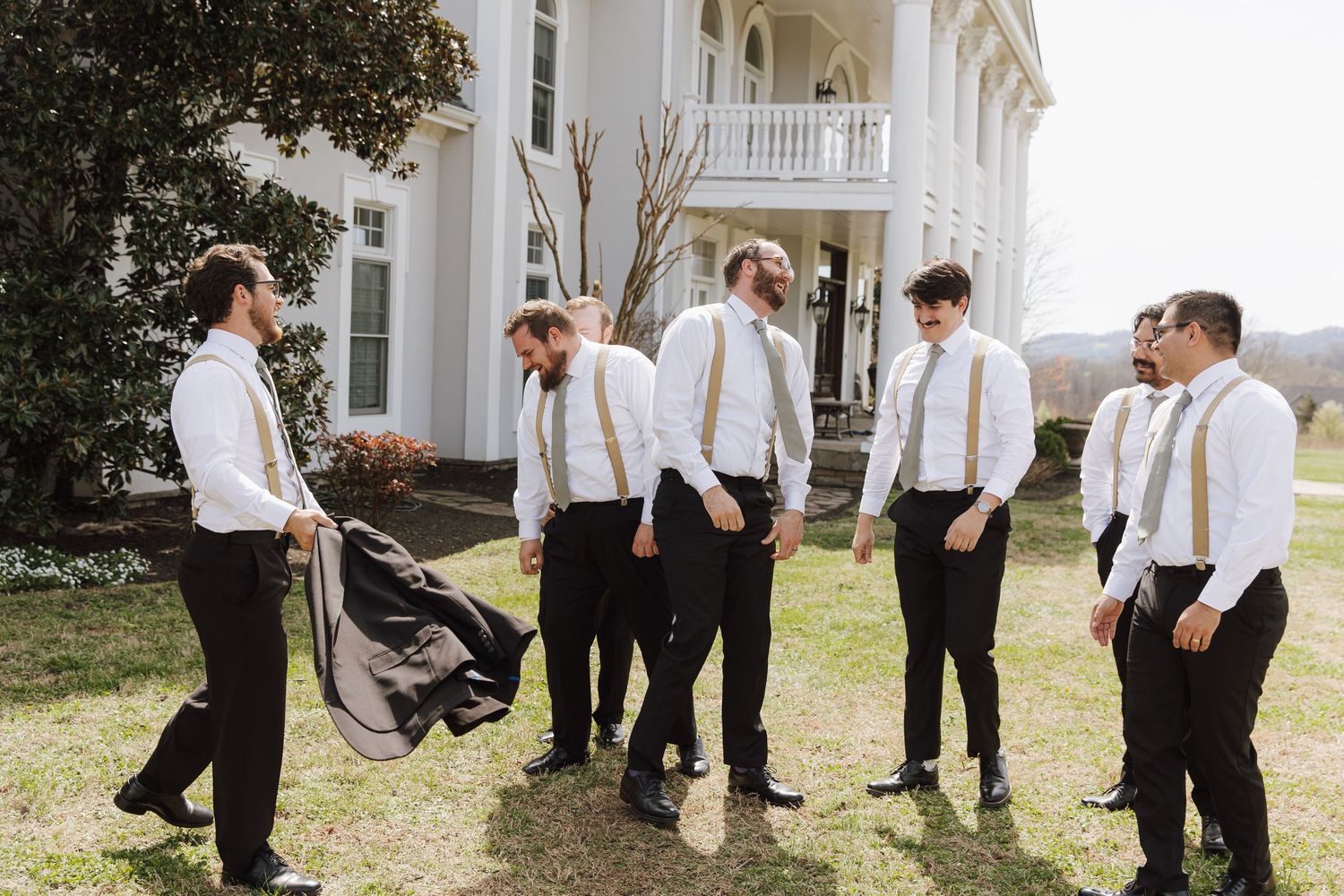 Groomsmen in white shirts, black pants, and gold suspenders gathered outside a white Southern mansion.