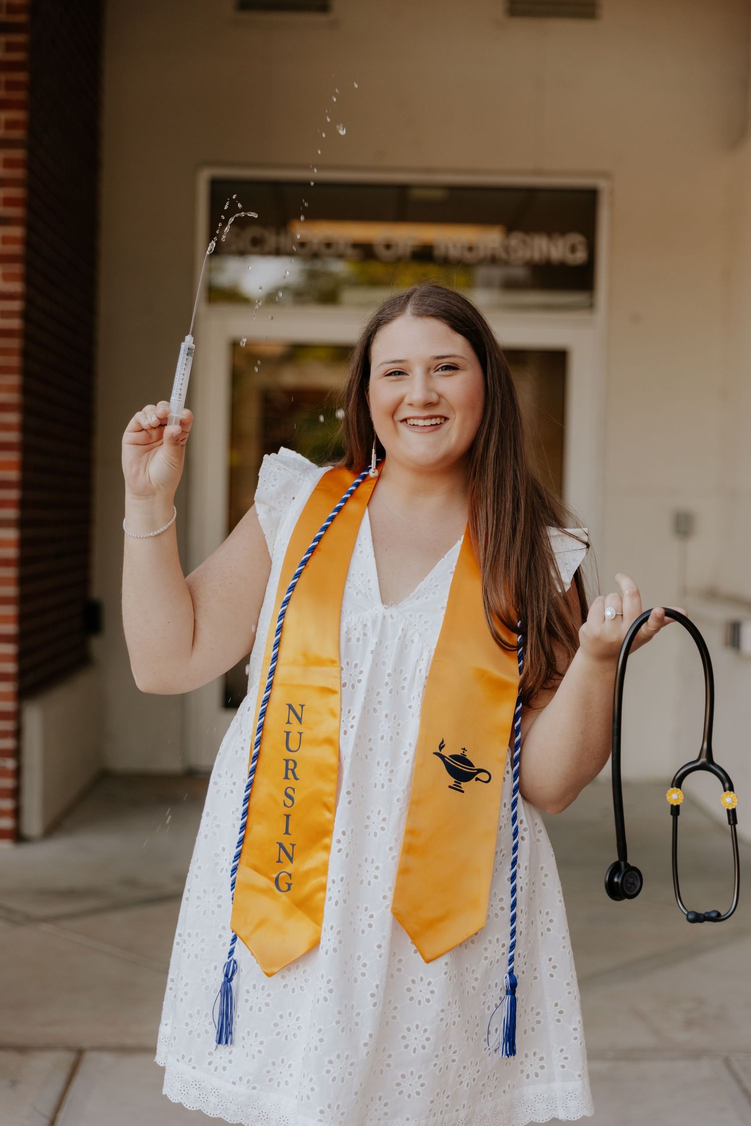 Graduate in white eyelet dress and yellow stole with blue tassel poses with diploma and stethoscope for graduation photos.