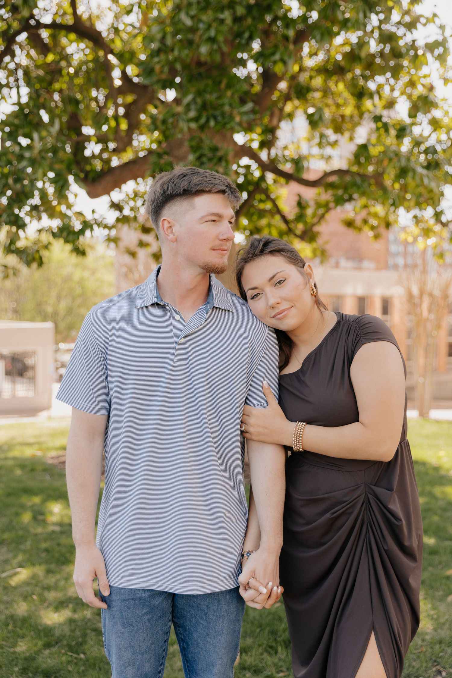 A couple stands close together outdoors on a sunny day, with green trees and a building in the background.