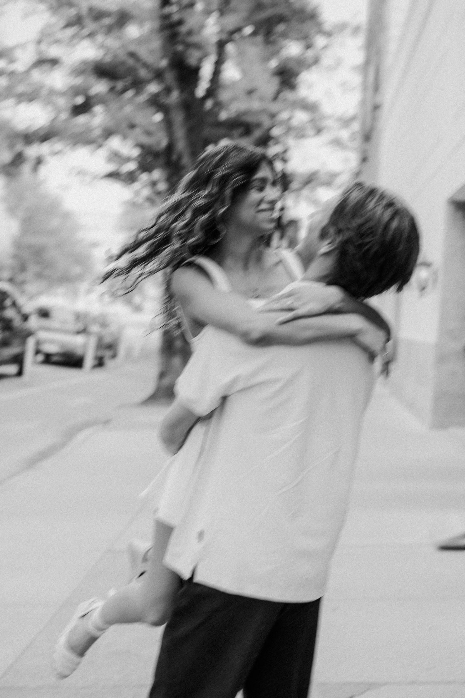 Black and white photo of a joyful couple spinning on a sidewalk, womans hair flowing as man lifts her.