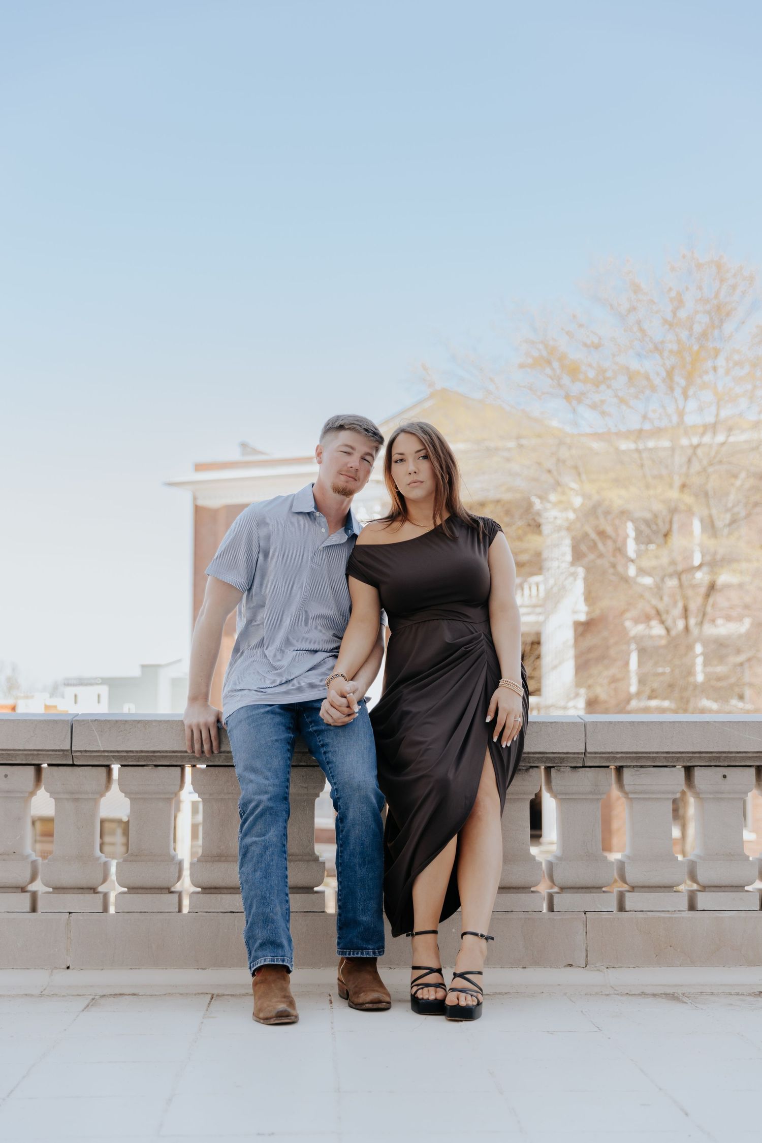 A couple stands together on a balcony with stone railings against a light blue sky and brick building backdrop.