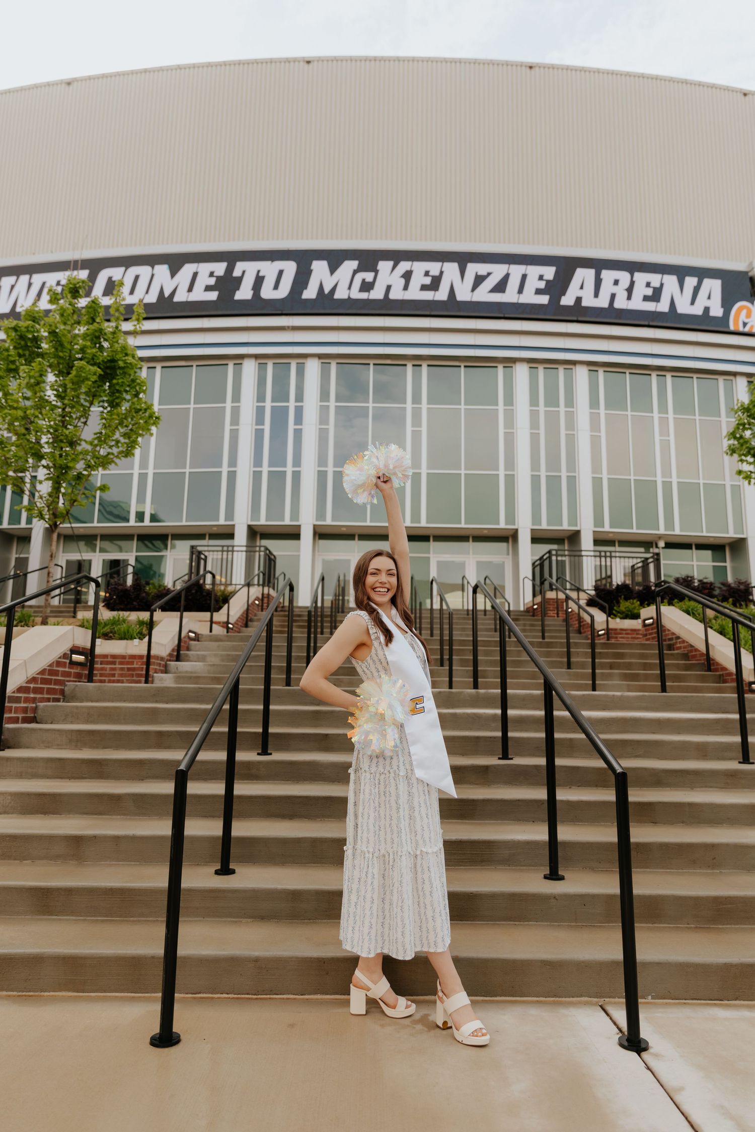 A person in a white dress poses on the steps outside McKenzie Arena on a sunny day.