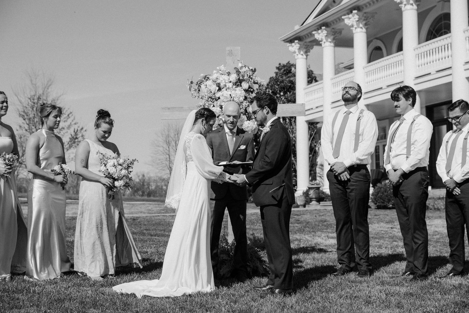Black and white outdoor wedding ceremony with bride, groom, officiant, and wedding party at a historic estate.