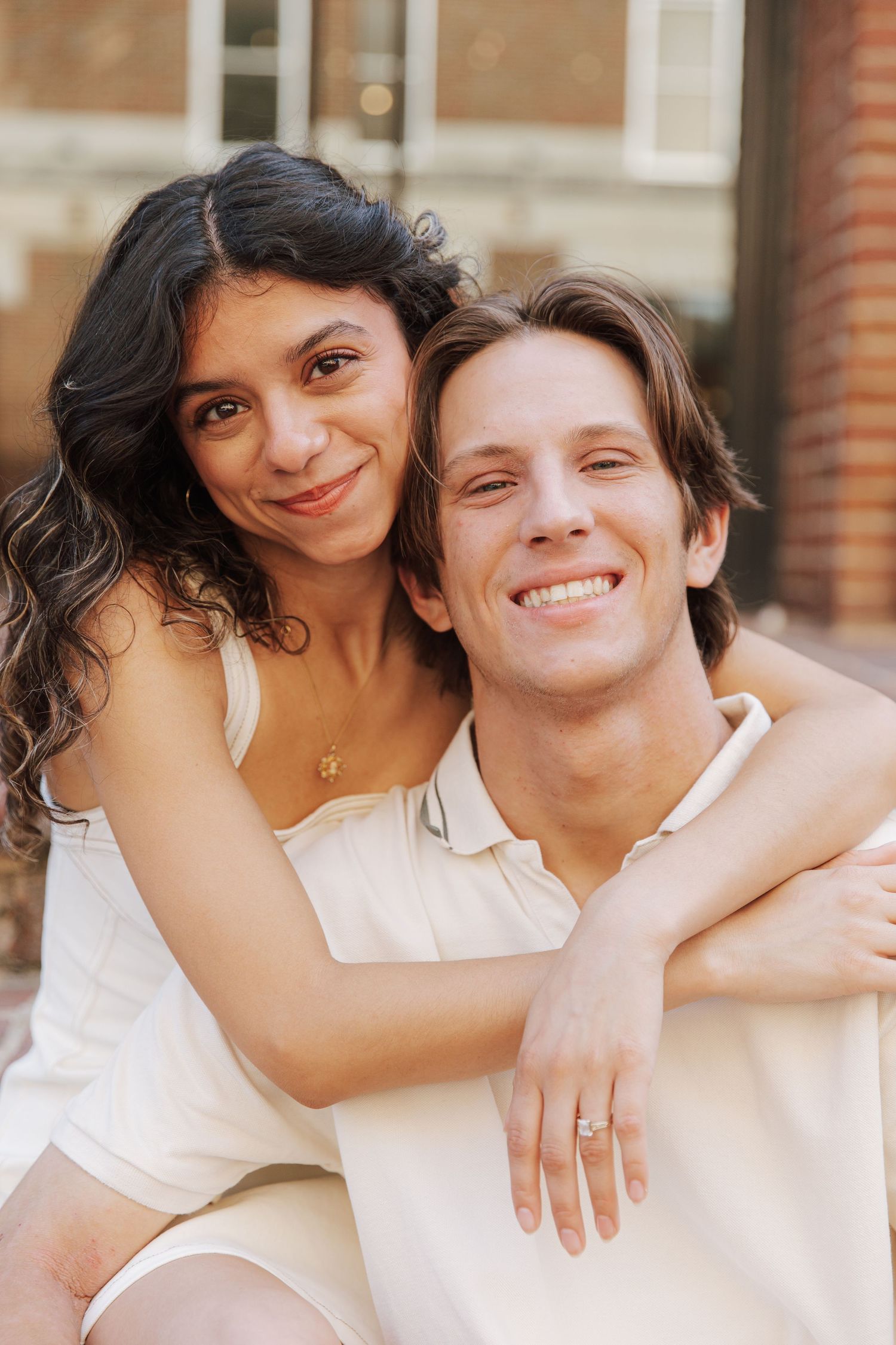 A smiling couple in cream outfits embrace outdoors, woman wrapping arms around mans shoulders in a warm pose.