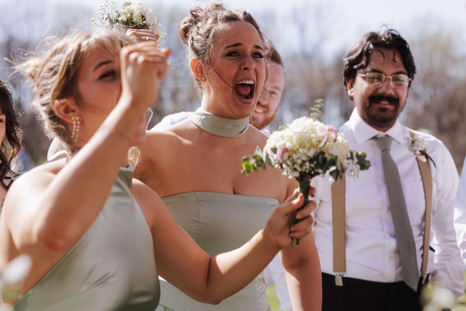 Joyful bridesmaids in sage green dresses celebrating outdoors holding white bouquets at a wedding.