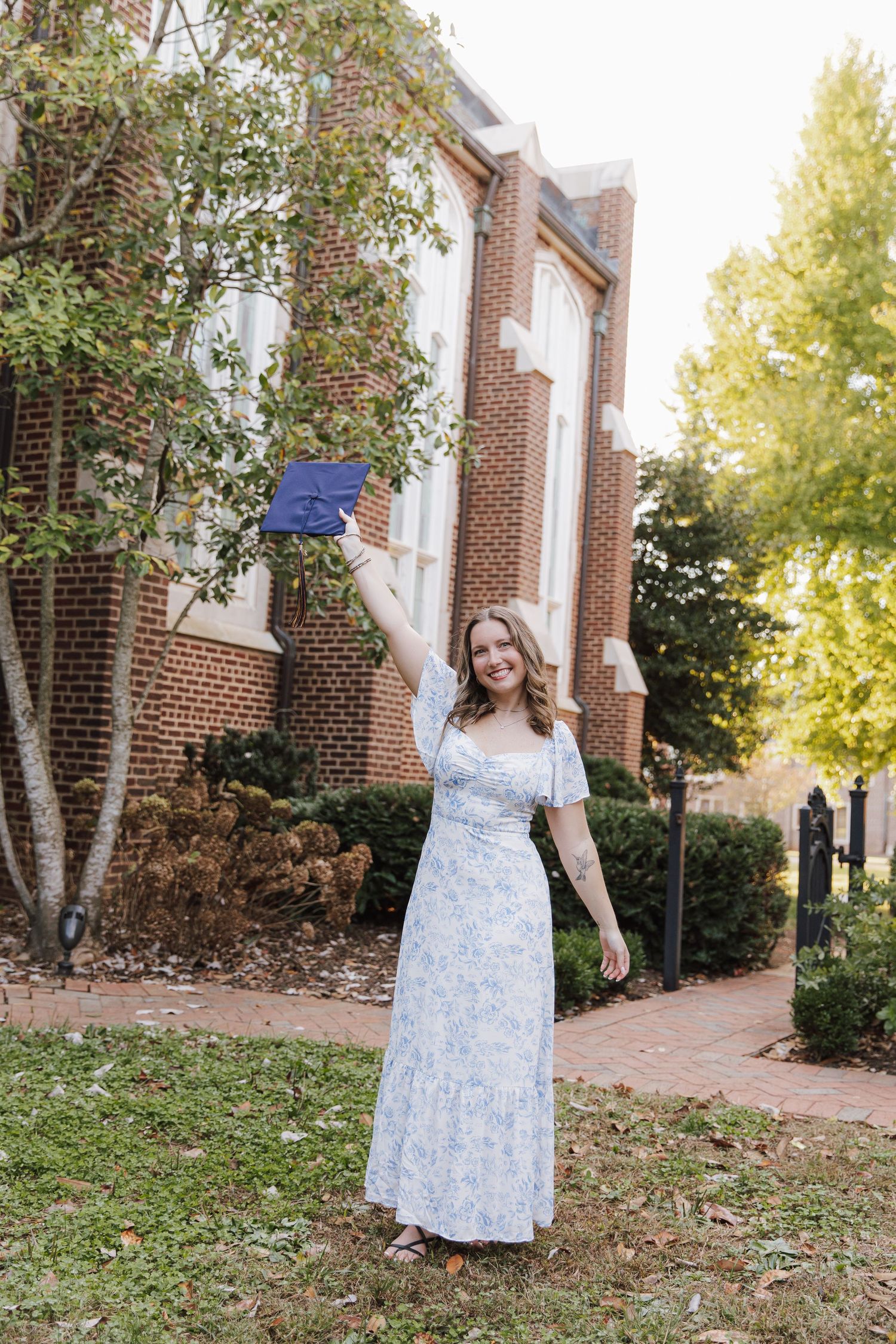 Graduate in a light blue floral dress poses outdoors near brick buildings during autumn.