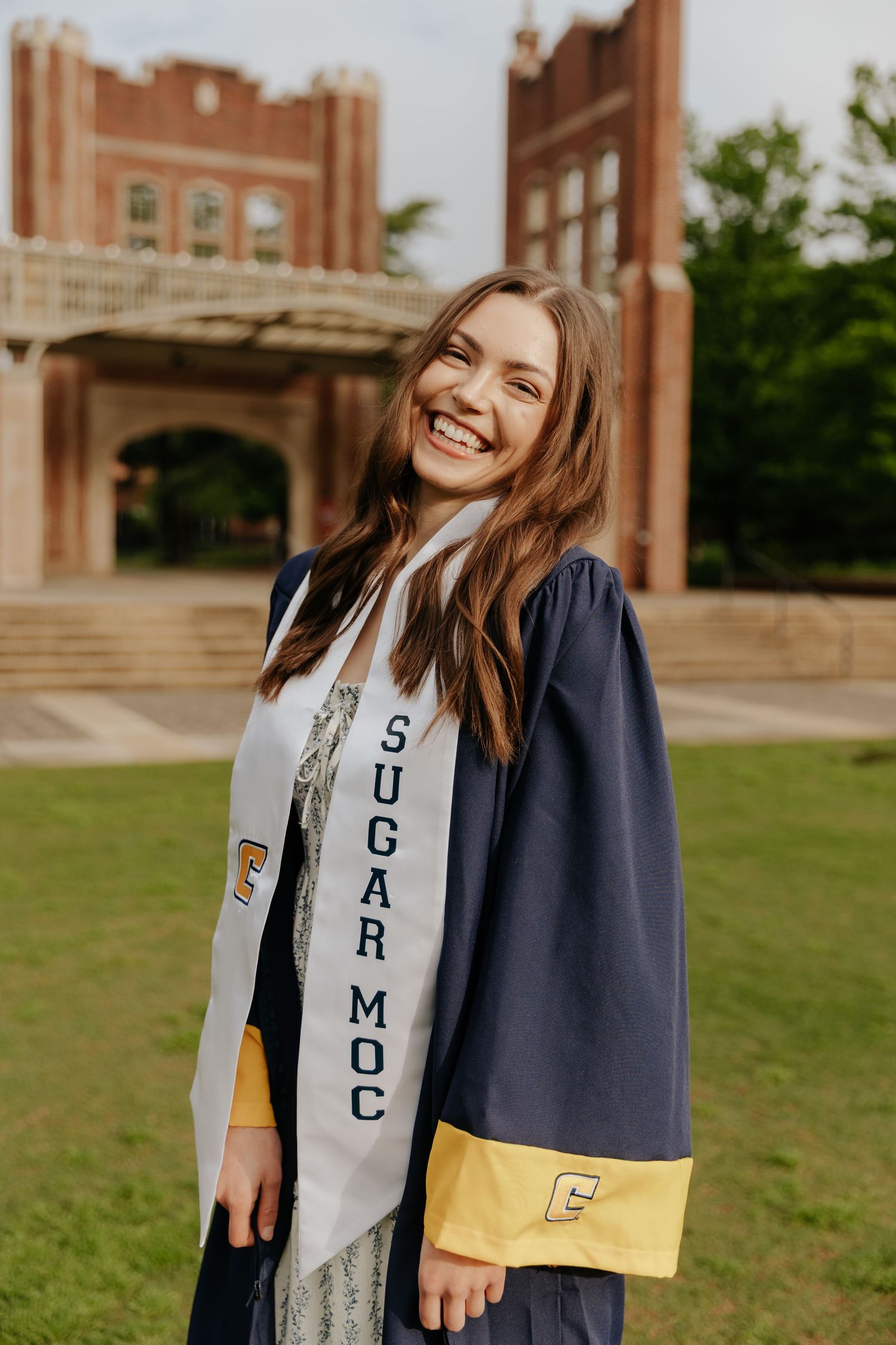 Graduate in navy and gold academic regalia poses on campus with historic buildings in background.