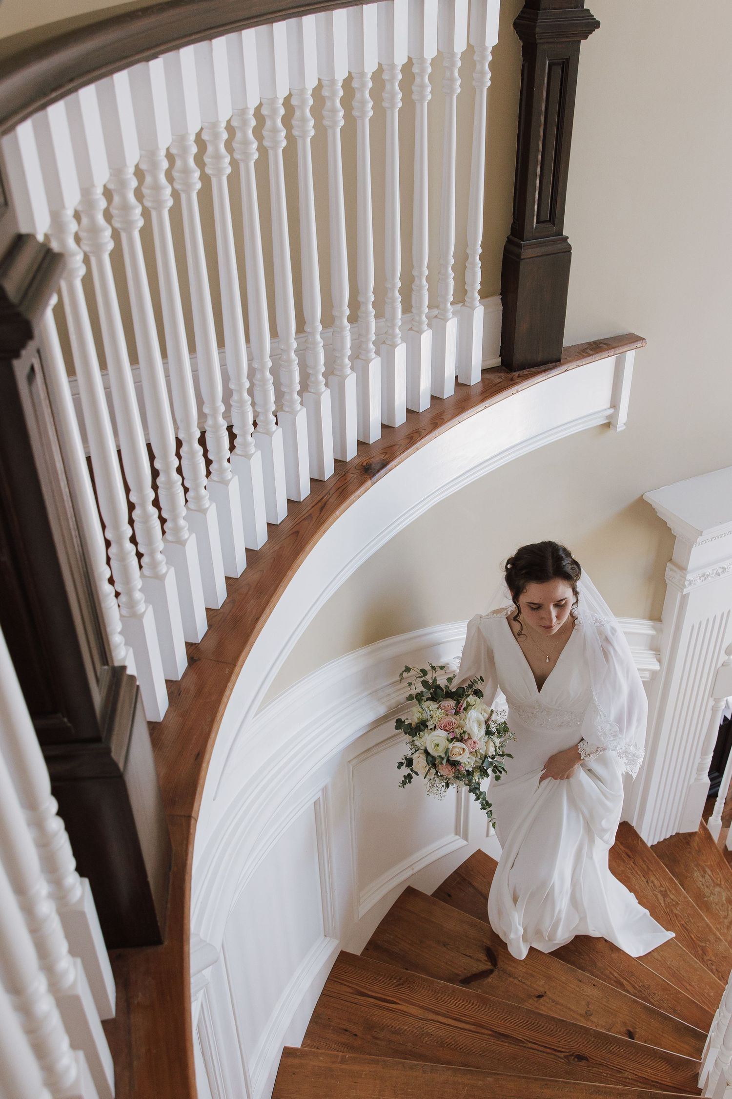 Aerial view of bride in white long-sleeve gown holding floral bouquet descending elegant curved wooden staircase.