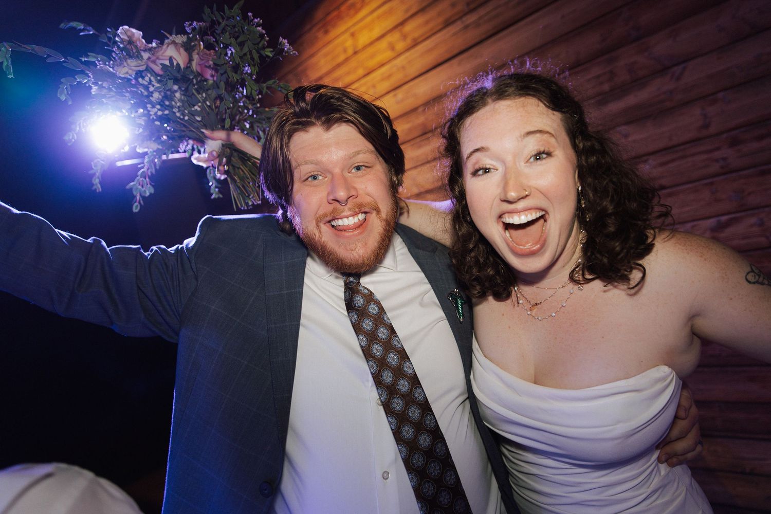 Joyful newlywed couple celebrating at their wedding reception, groom raising bouquet while bride laughs with excitement.