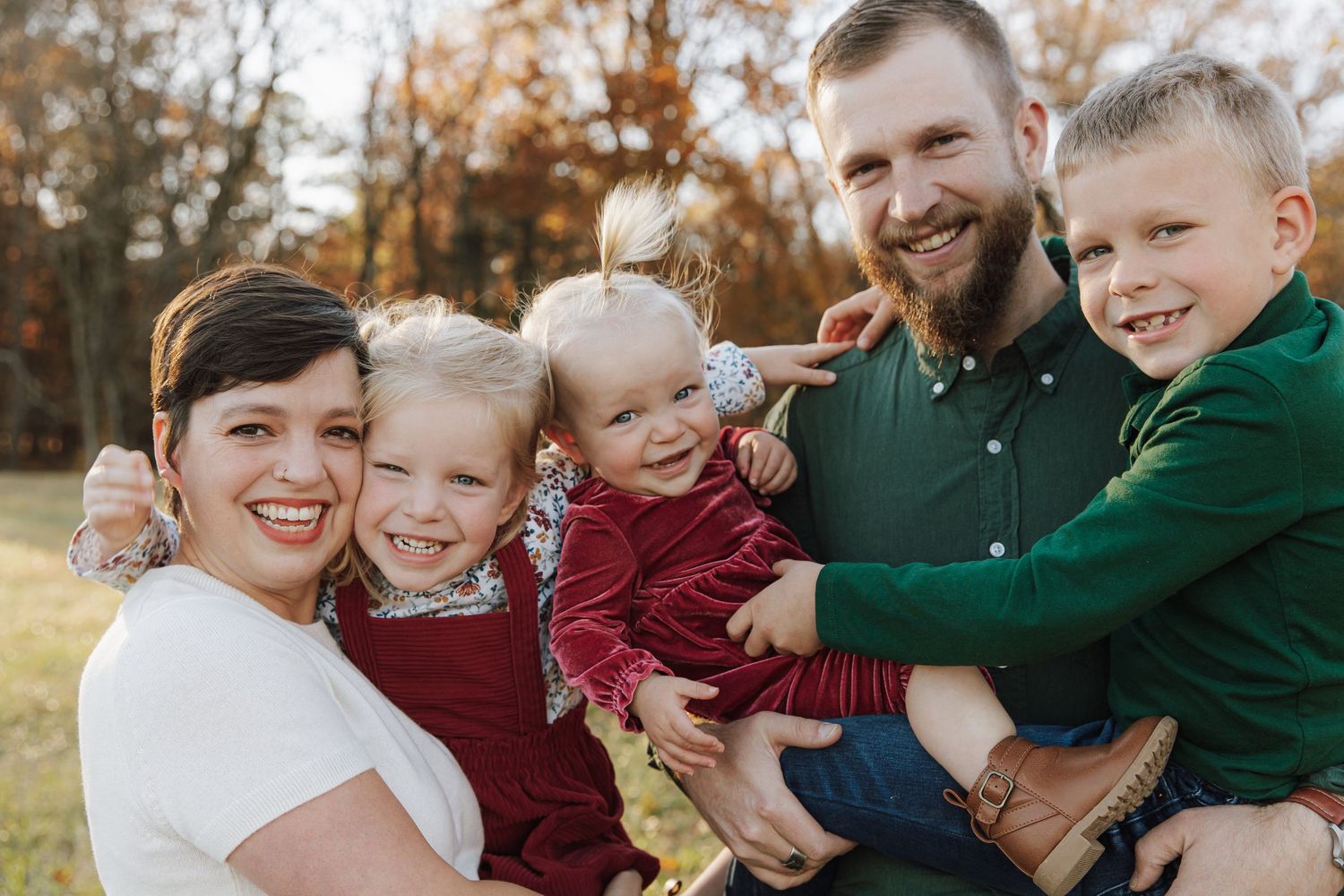 A joyful family portrait taken outdoors during autumn with fall foliage in the background.