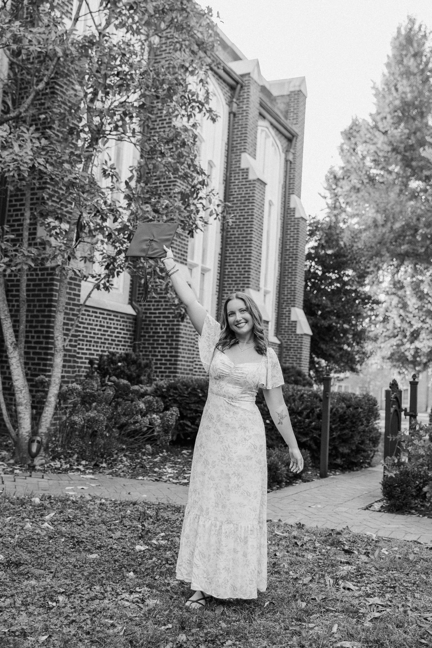 Graduate in white lace dress poses for portraits on lawn near brick building in black and white.
